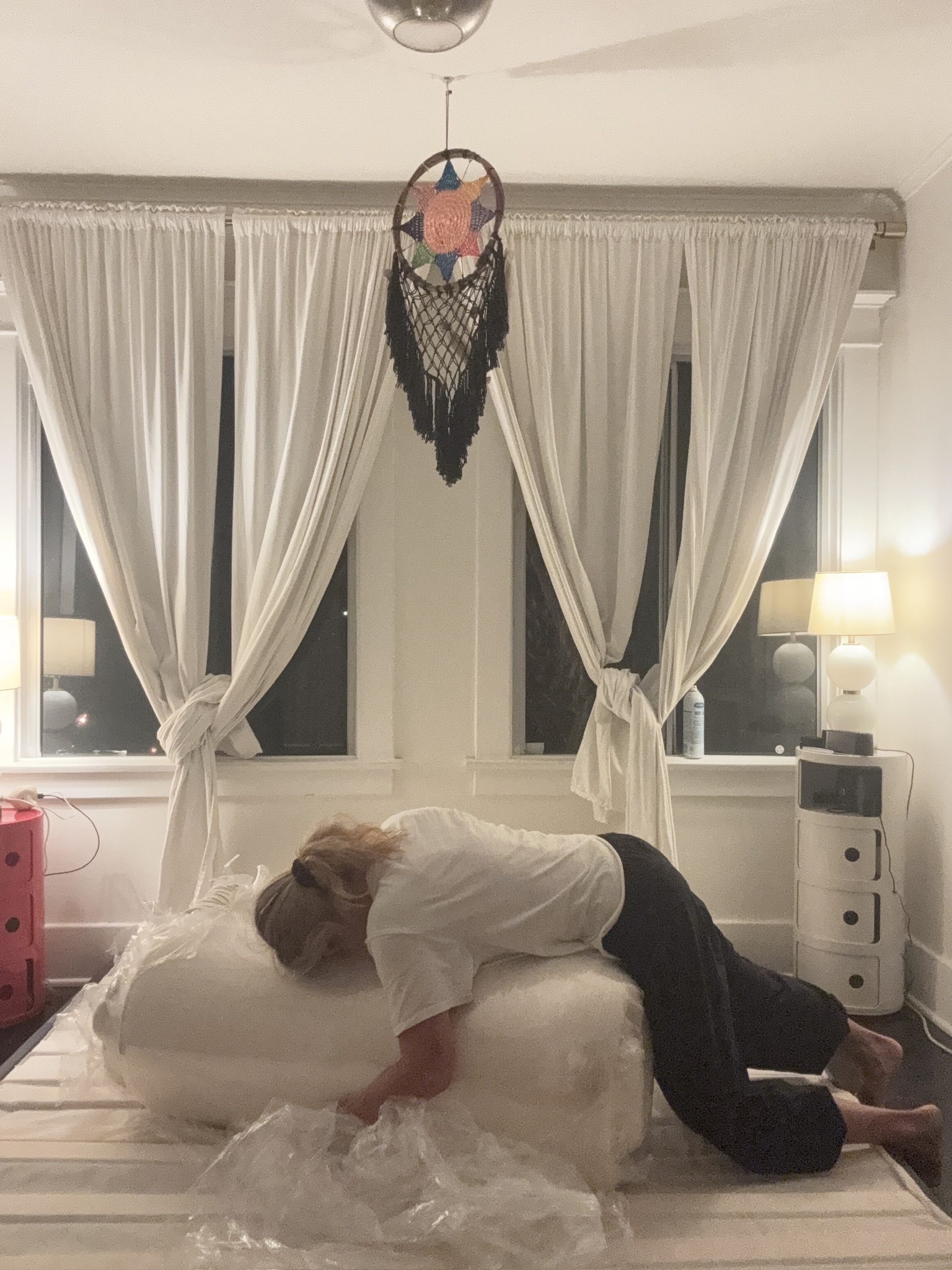 A person kneels on a bed, unrolling a mattress from plastic packaging in a softly lit bedroom with white curtains and a dreamcatcher hanging from the ceiling.