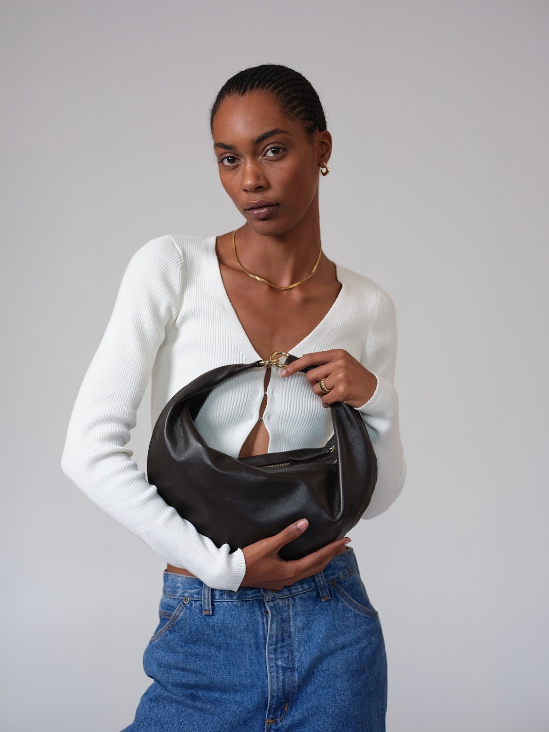A person in a white long-sleeve top and blue jeans holds a black crescent-shaped handbag against a plain background.