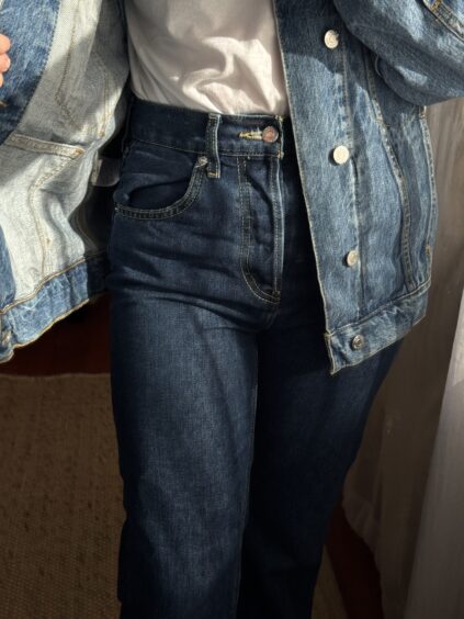 Person wearing a white T-shirt, blue denim jacket, and high-waisted blue jeans standing indoors with sunlight casting shadows.