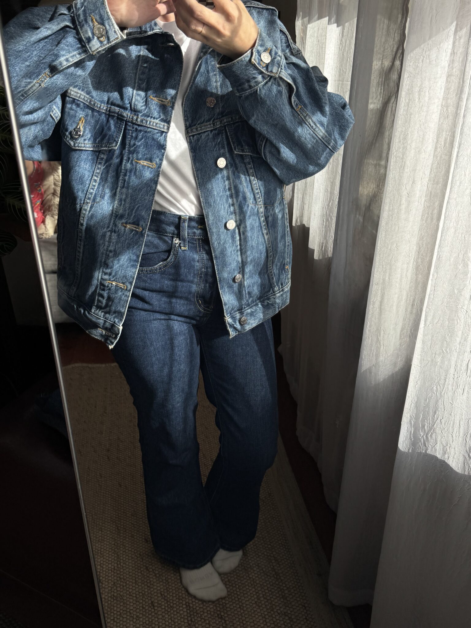 Person taking a mirror selfie wearing a blue denim jacket, white shirt, blue jeans, and white socks. Sunlight is streaming through sheer curtains.
