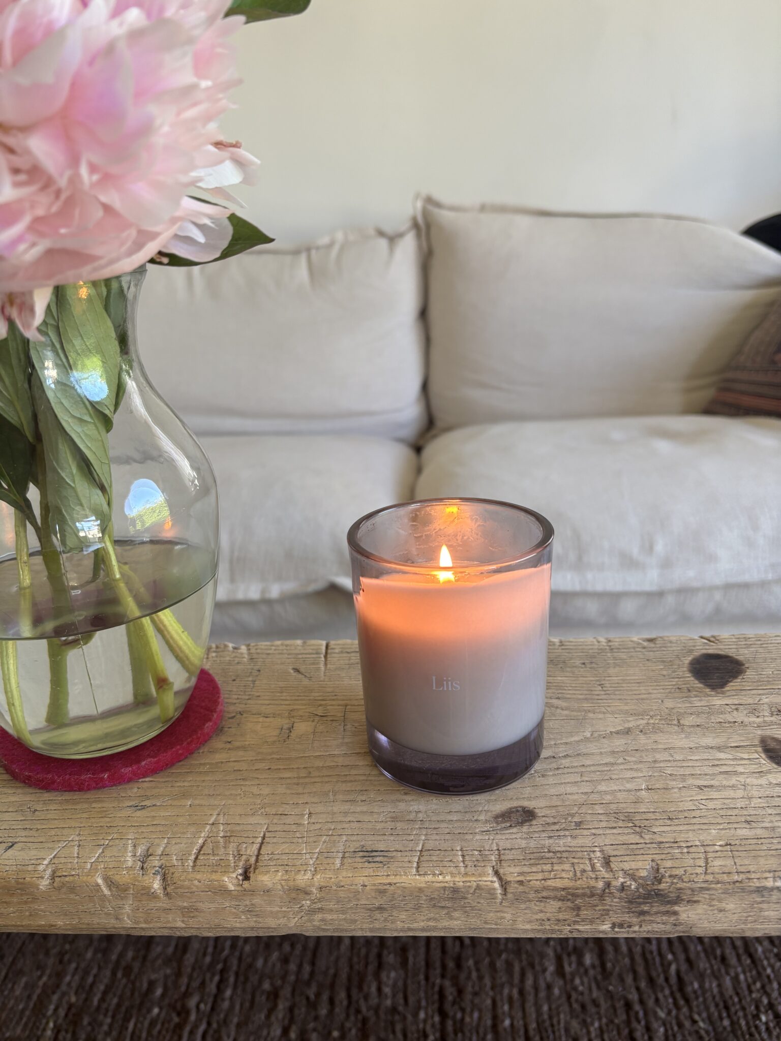A lit candle labeled "Lis" sits on a wooden table next to a vase of pink flowers, with a beige sofa in the background.