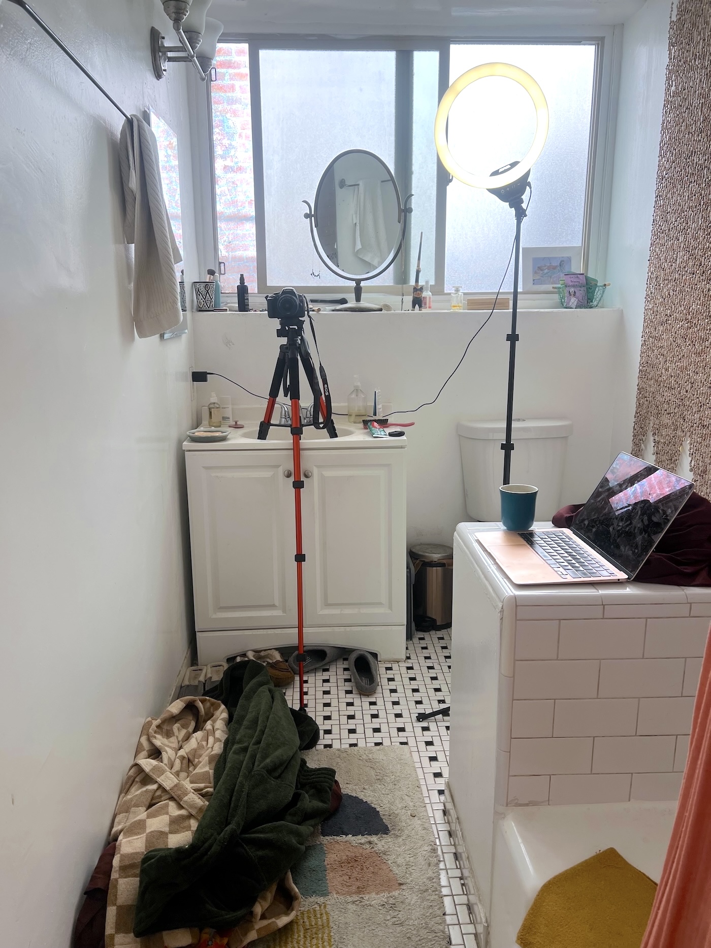 A bathroom with a tripod camera, ring light, and laptop set up near a sink; towels and blankets are on the floor.