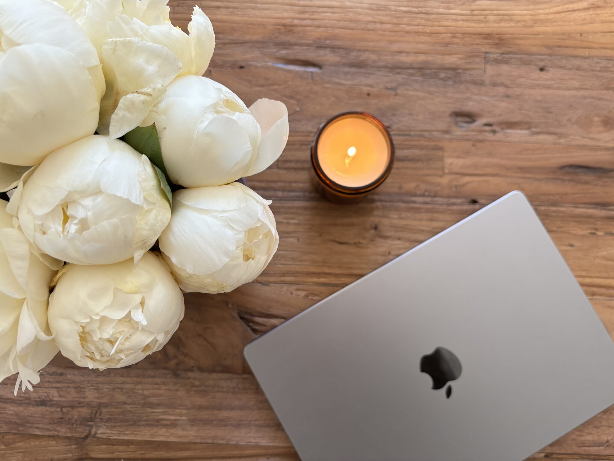 A closed silver MacBook, a lit candle, and a bouquet of white flowers are arranged on a wooden surface.