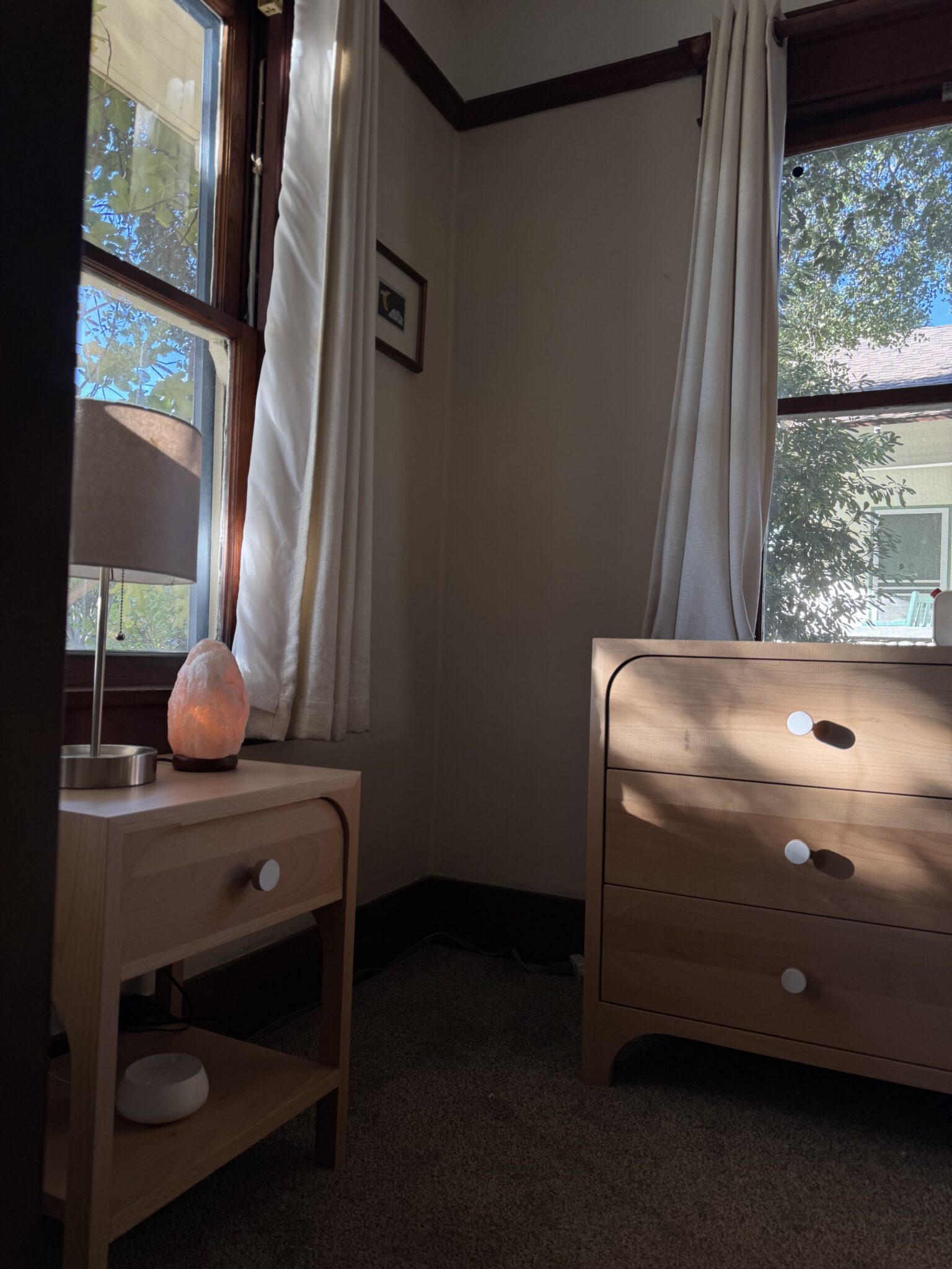 A sunlit corner of a room with a wooden dresser, nightstand holding a lamp and salt lamp, white curtains, and large windows showing trees outside.