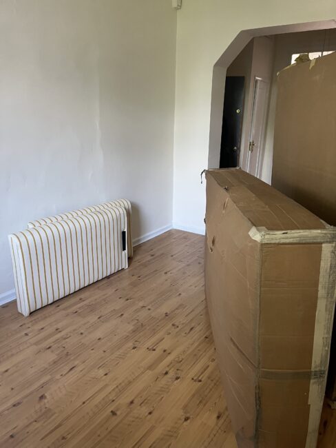 A room with a wooden floor, a striped cushion or mattress leaning against the wall, and a large cardboard box standing upright near the center.