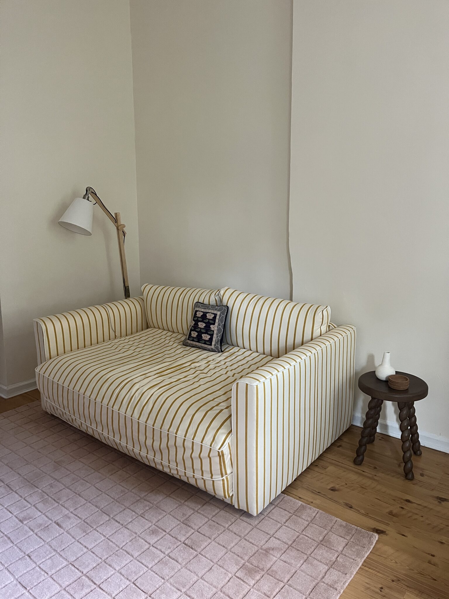 A yellow and white striped sofa with a patterned cushion sits next to a wooden side table and floor lamp on a pink rug in a room with light-colored walls and wooden floor.