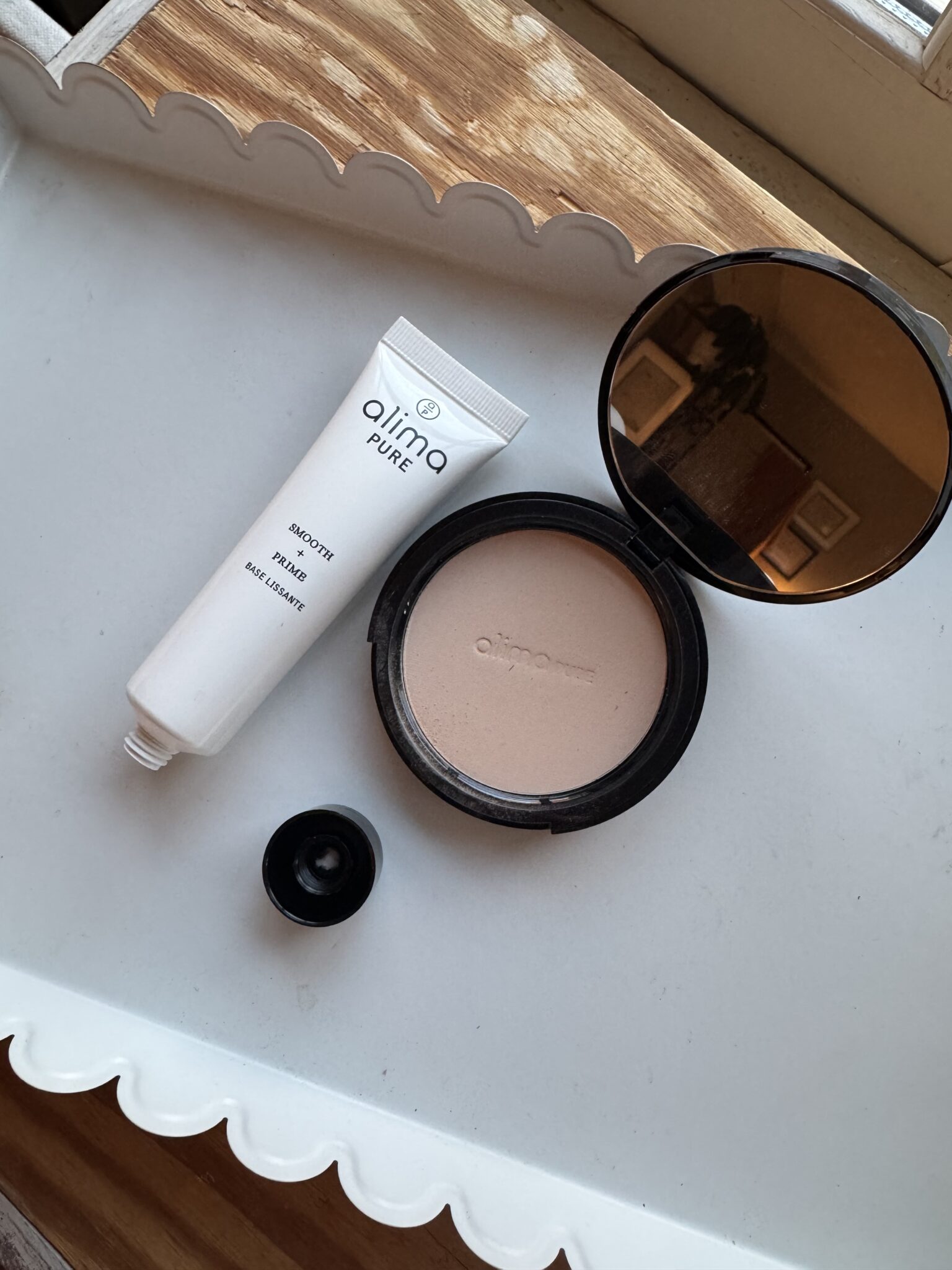 A tube of Alima Pure Smooth + Prime, an open container of pressed powder with mirror, and a black cap on a white tray near a window.