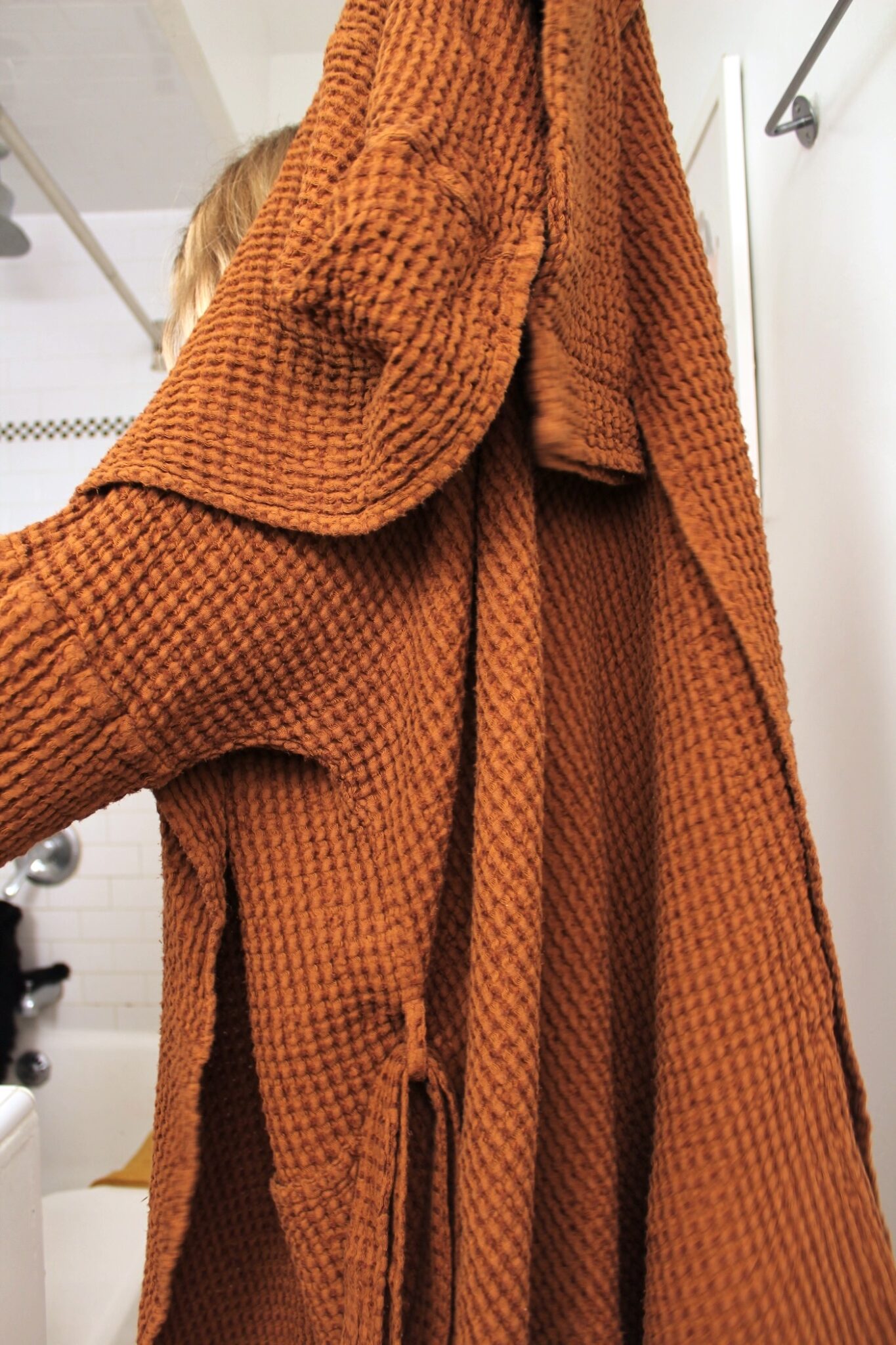 A person holds up a textured, brown robe in a bathroom with white tiles visible in the background.