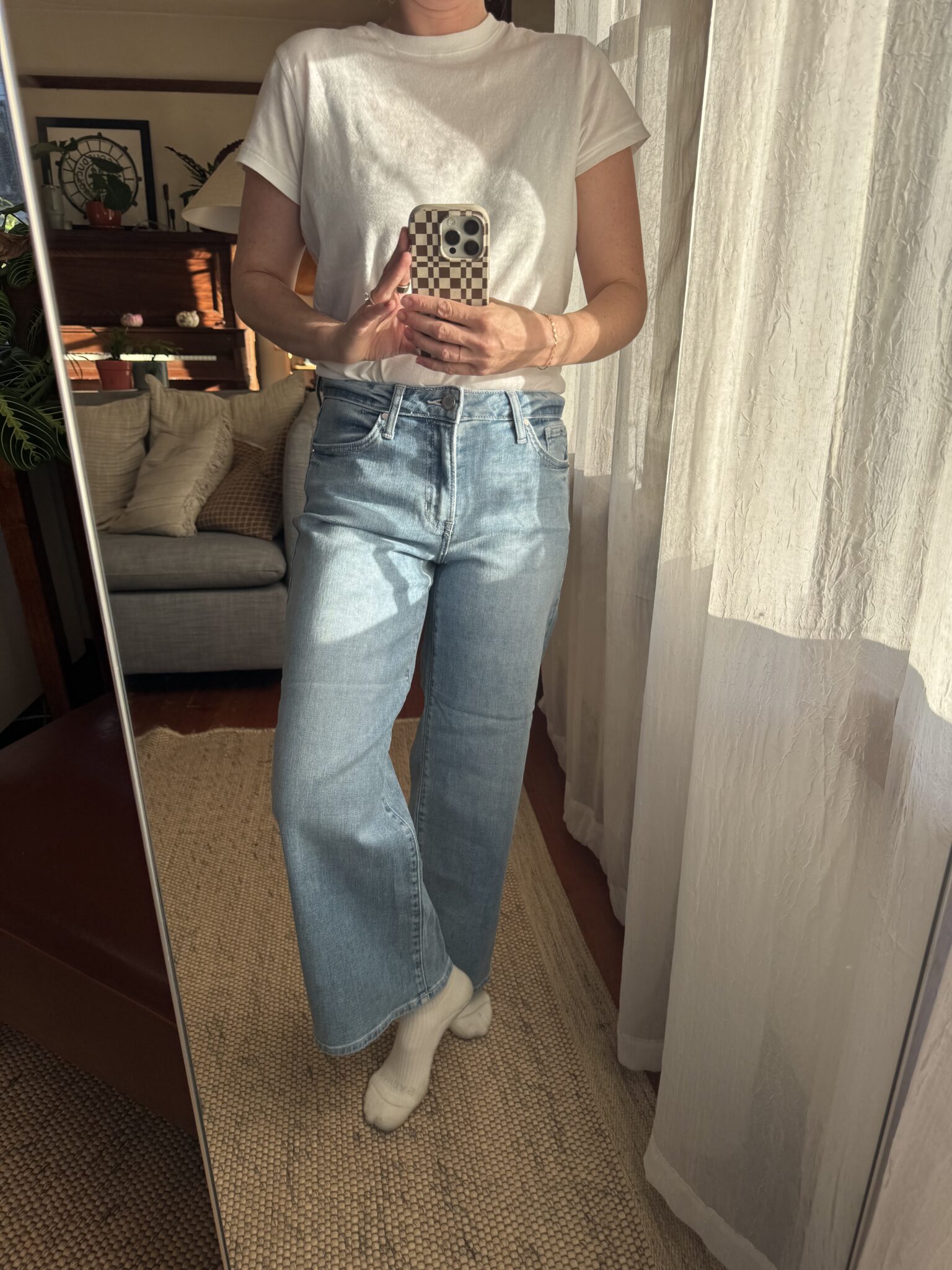 Person wearing a white t-shirt, light blue wide-leg jeans, and white socks stands indoors, taking a mirror selfie with a checkered phone case.