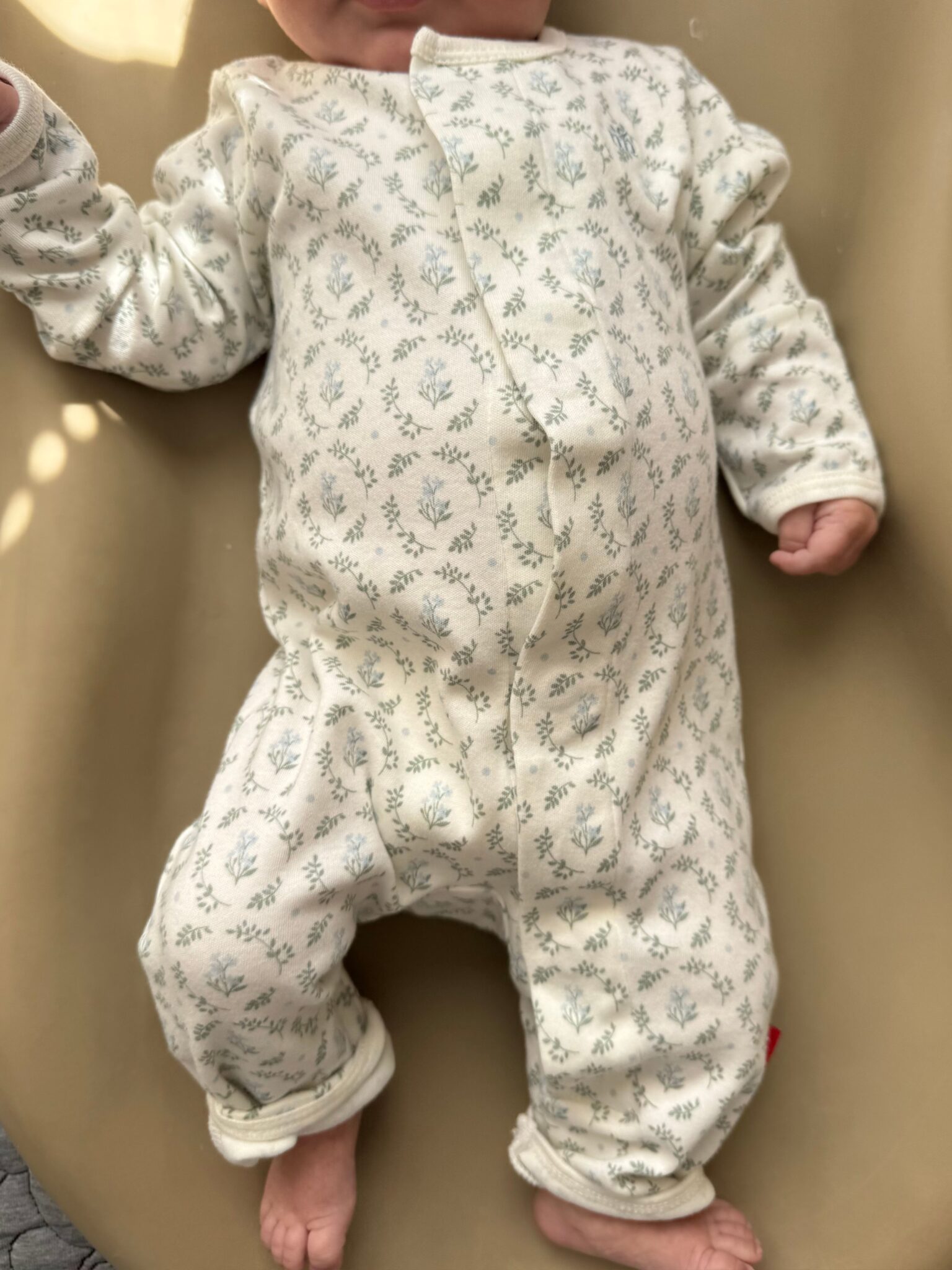 A baby wearing a long-sleeved, white onesie with a small blue floral pattern, lying on a beige surface.