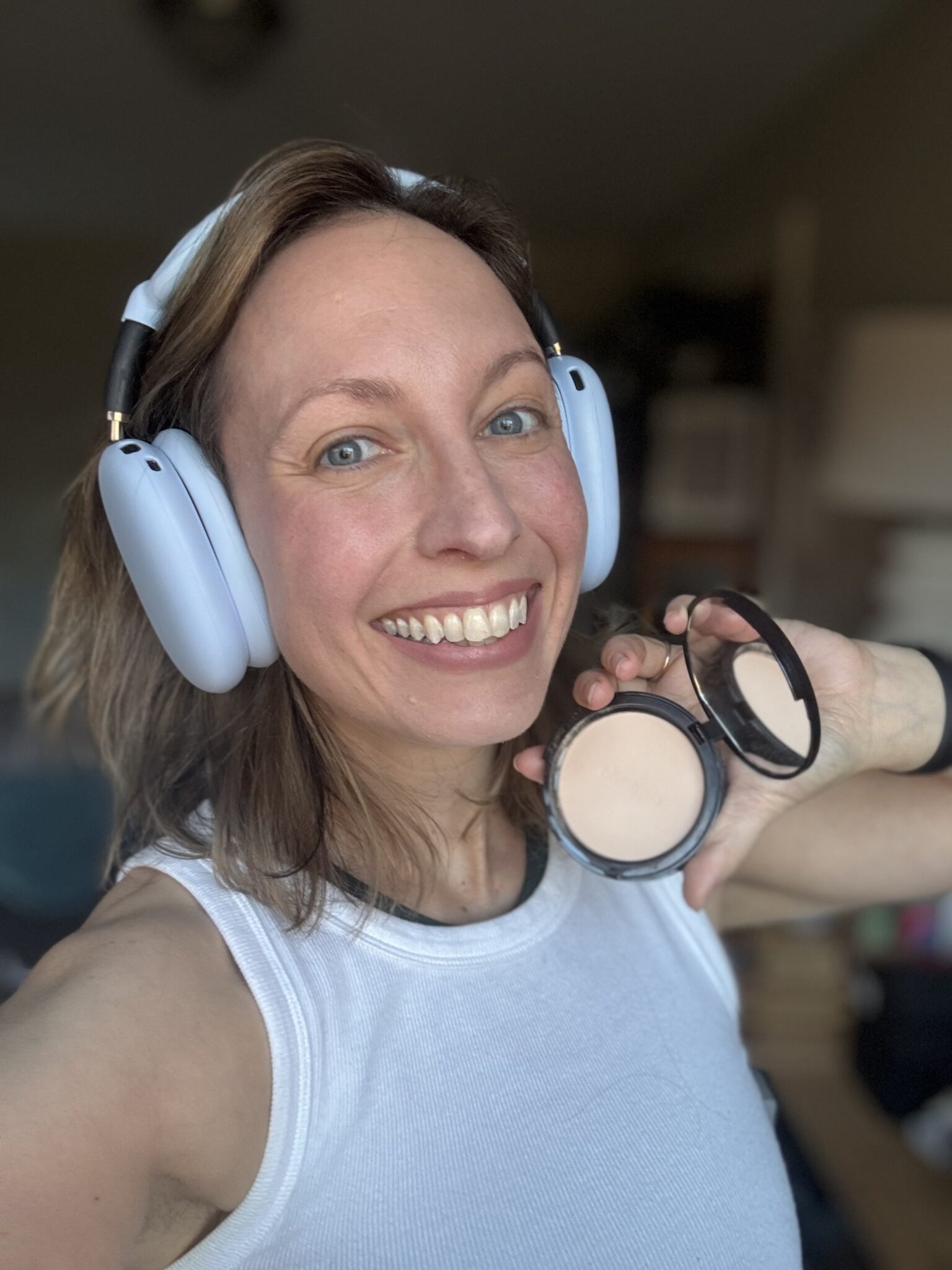 Woman wearing headphones and a white tank top smiles at the camera, holding an open compact of pressed powder makeup.