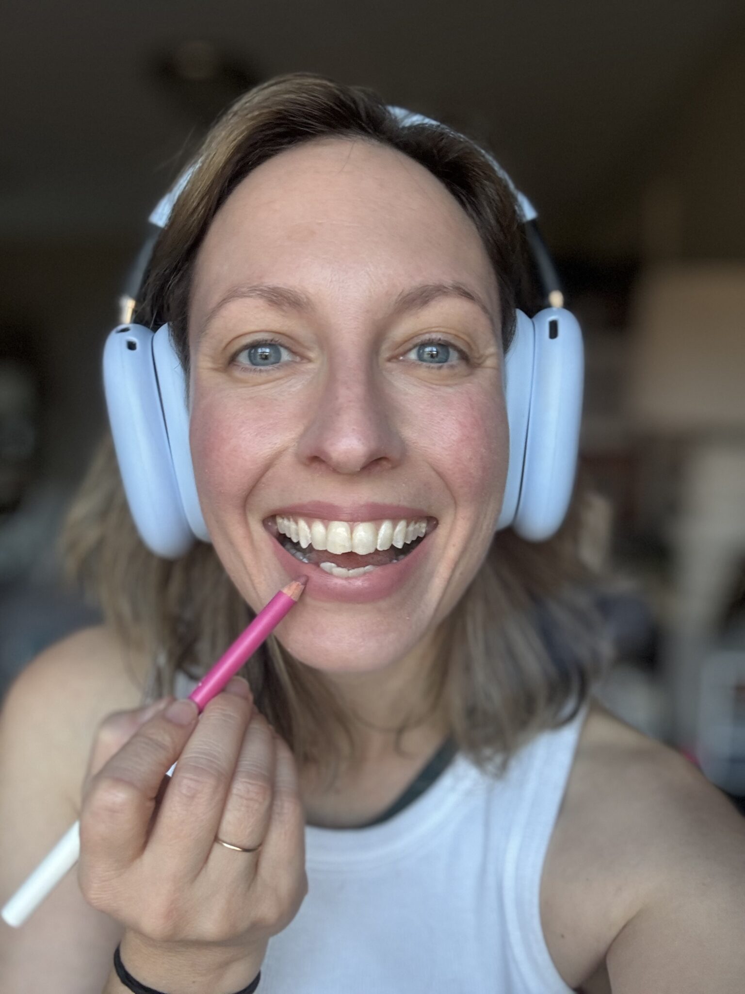 A person wearing white headphones and a white tank top applies pink lip liner while smiling at the camera.