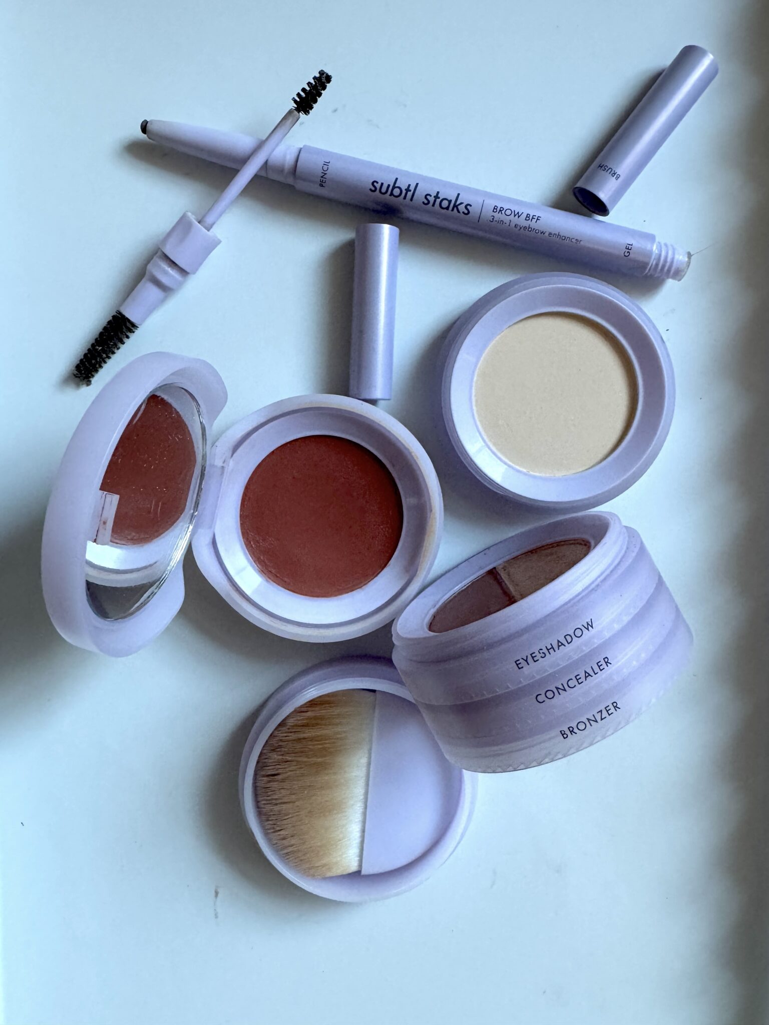 A flat lay of purple cosmetic containers including brow kit, cream blush, powder, and a makeup brush labeled eyeshadow, concealer, and bronzer on a white background.