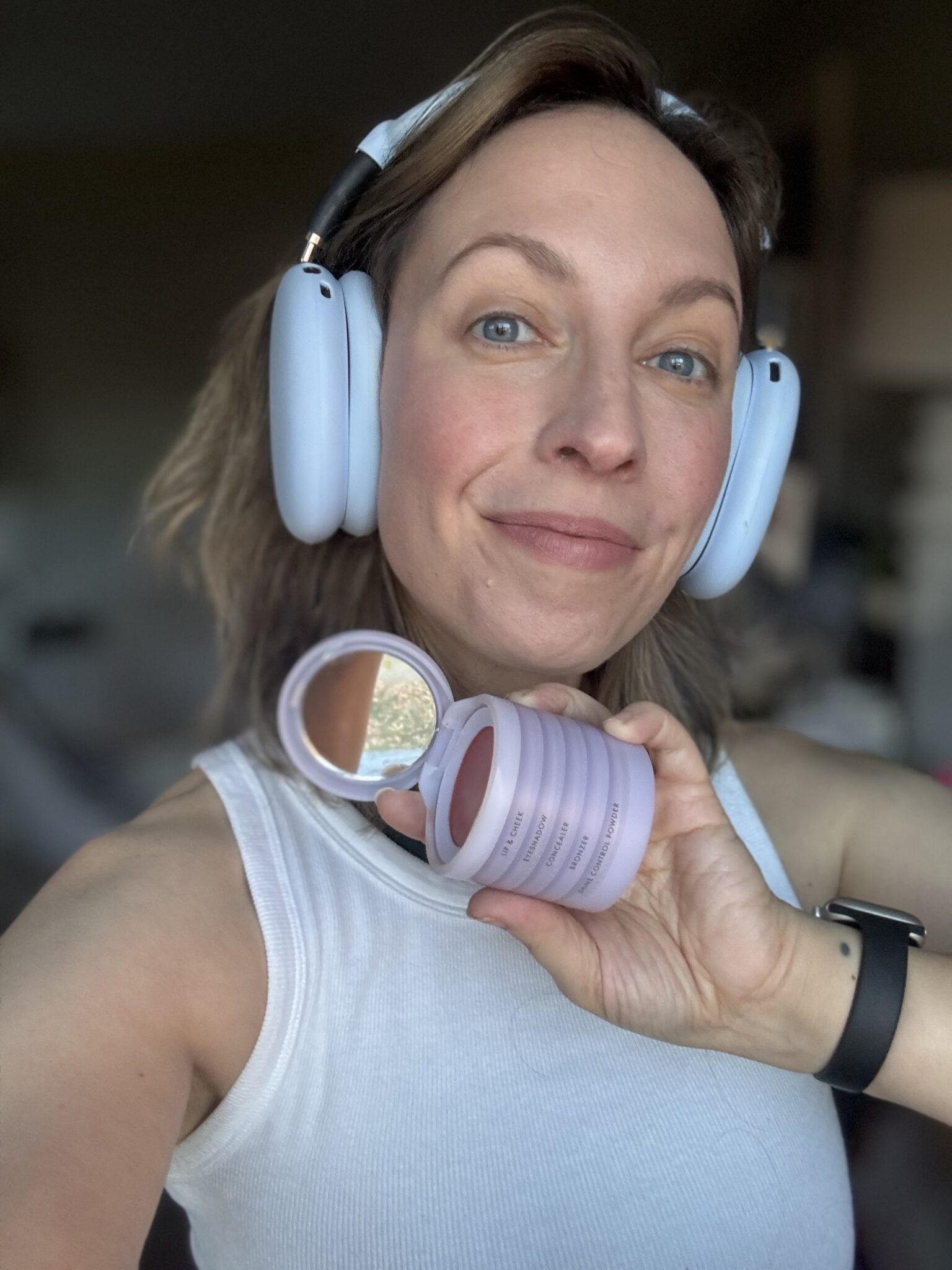 A person wearing headphones and a white tank top holds an open compact makeup container with a mirror, smiling at the camera.