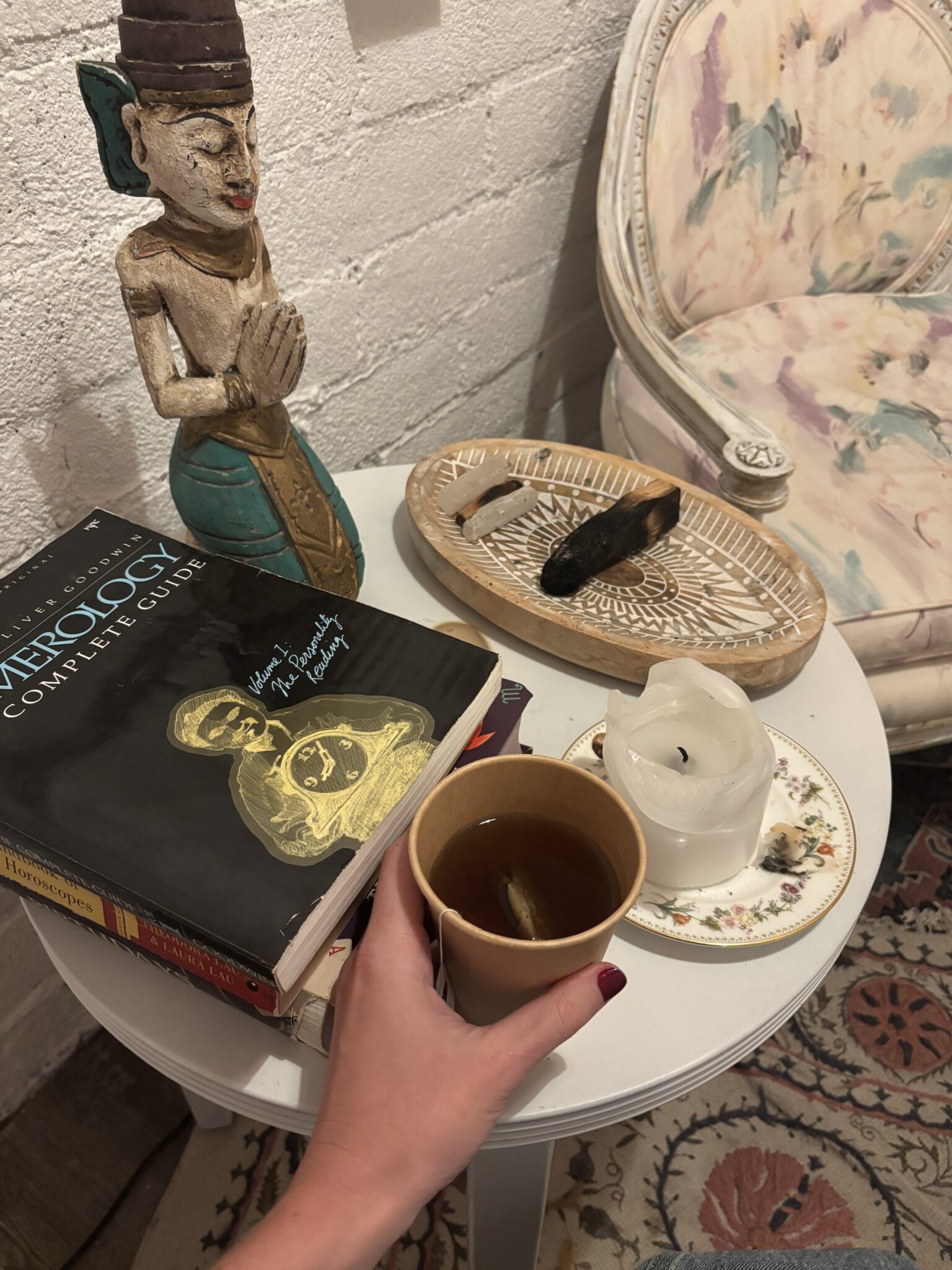 A hand holds a cup of tea over a small table with a numerology book, a white candle, an incense stick, a wooden statue, and a teacup and saucer beside a patterned armchair.