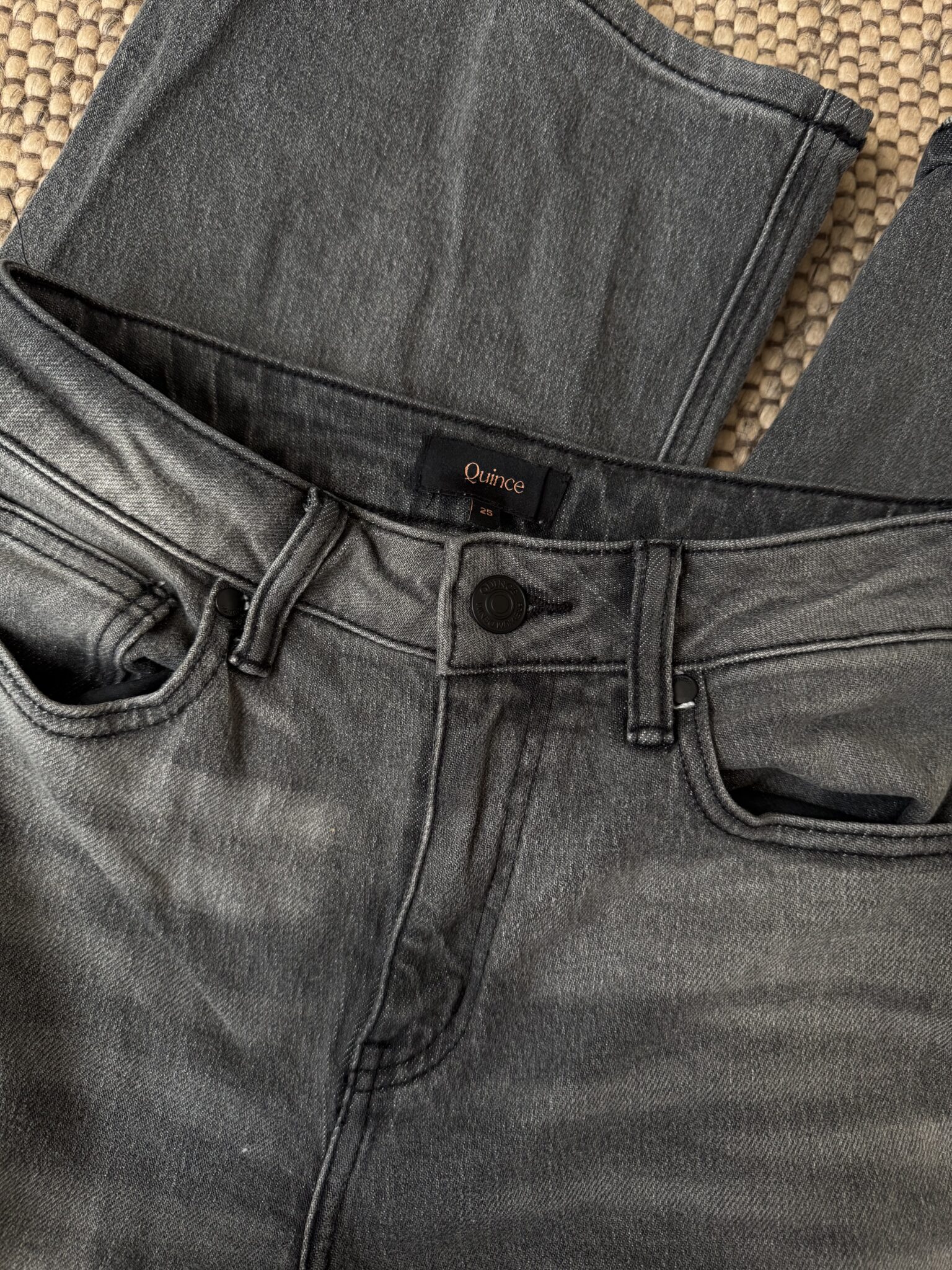A pair of faded black jeans with a visible Quince brand label, laid flat on a textured beige surface.