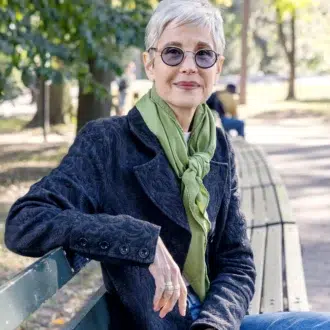 An older woman with short gray hair, glasses, and a green scarf sits on a park bench outdoors, looking at the camera.