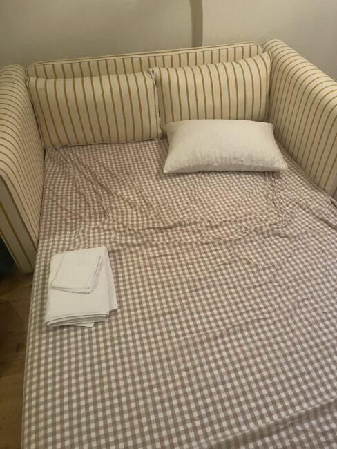 A daybed with yellow and white striped cushions, topped with a white pillow and folded white towels on a brown checkered sheet.