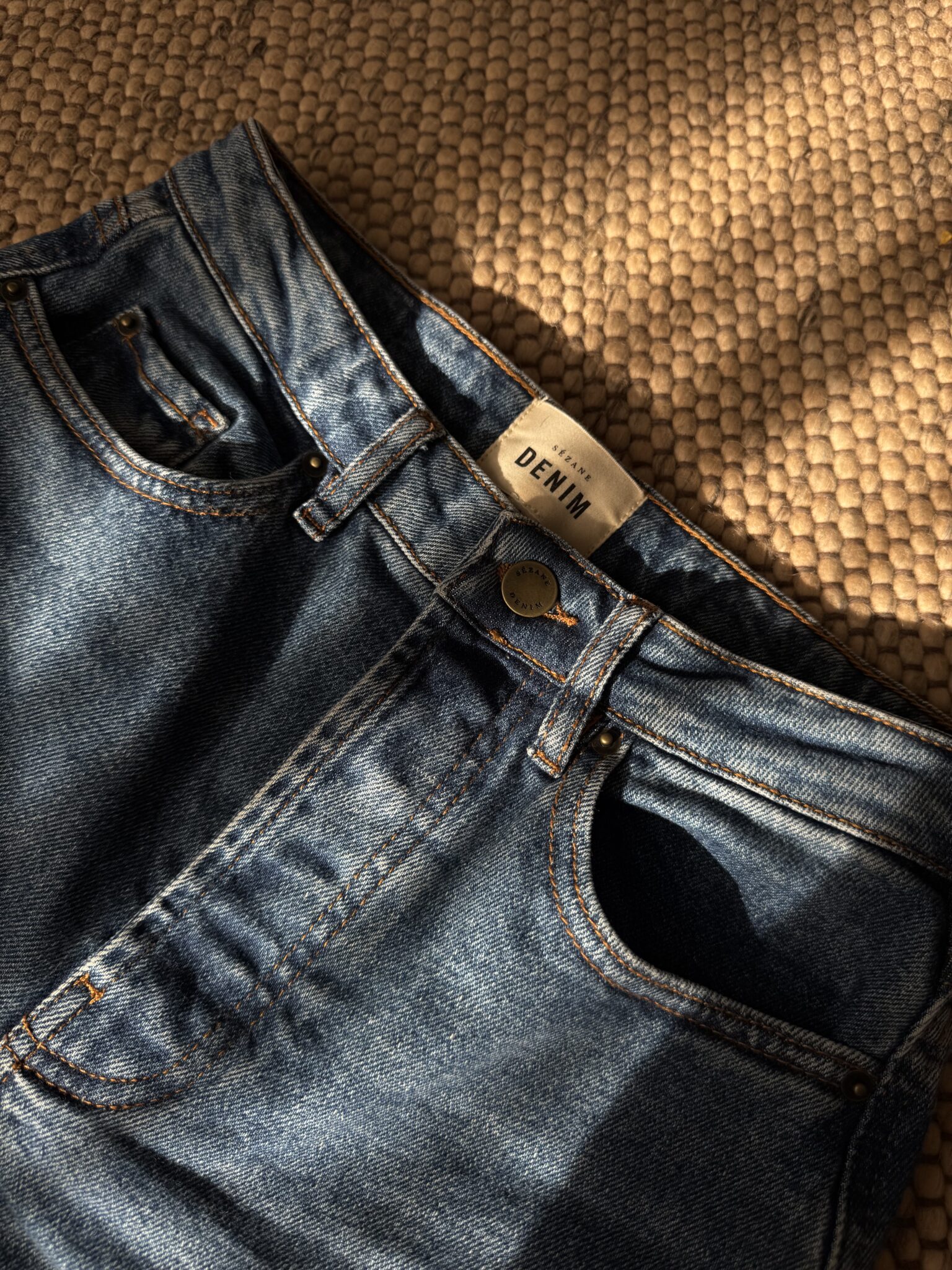 A pair of blue denim jeans is lying on a textured beige surface, with sunlight casting shadows across the fabric.