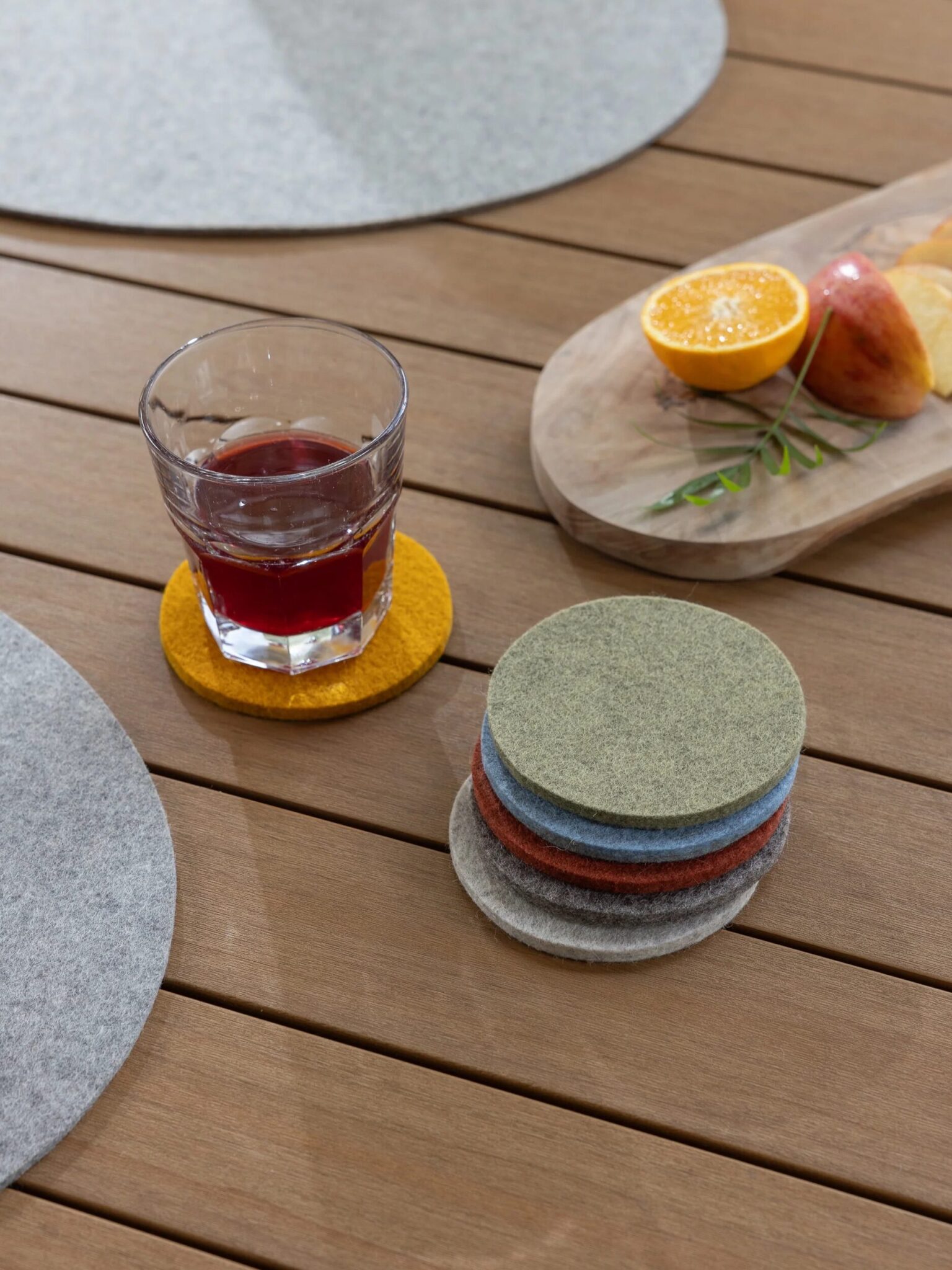 A glass of red beverage on a yellow felt coaster sits on a wooden table next to a stack of colorful coasters and a tray with sliced fruit.