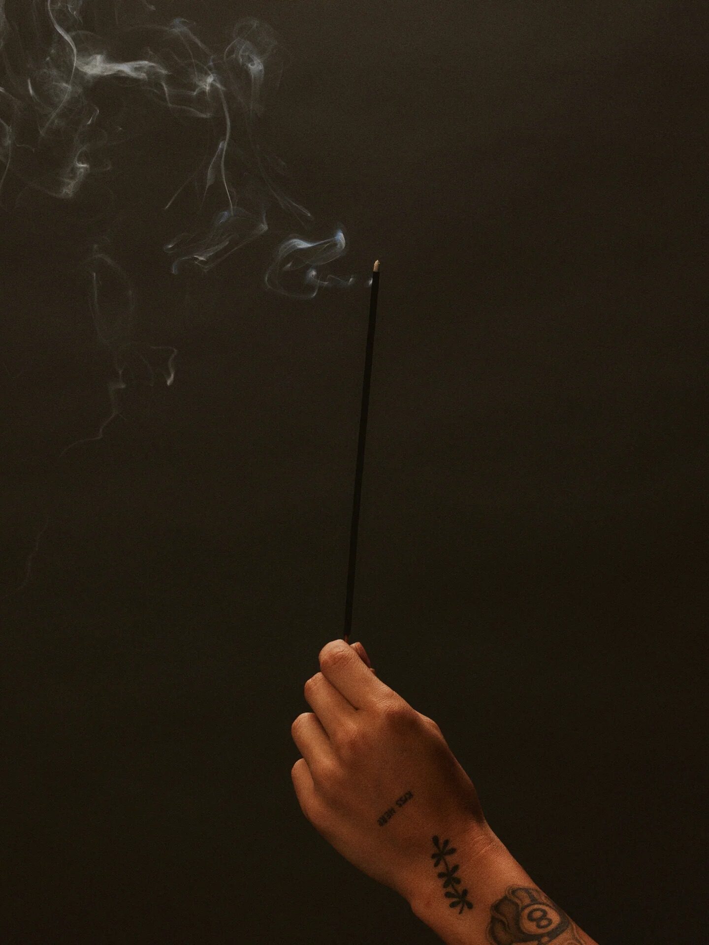 A hand with tattoos holds a lit incense stick, with smoke rising against a dark background.