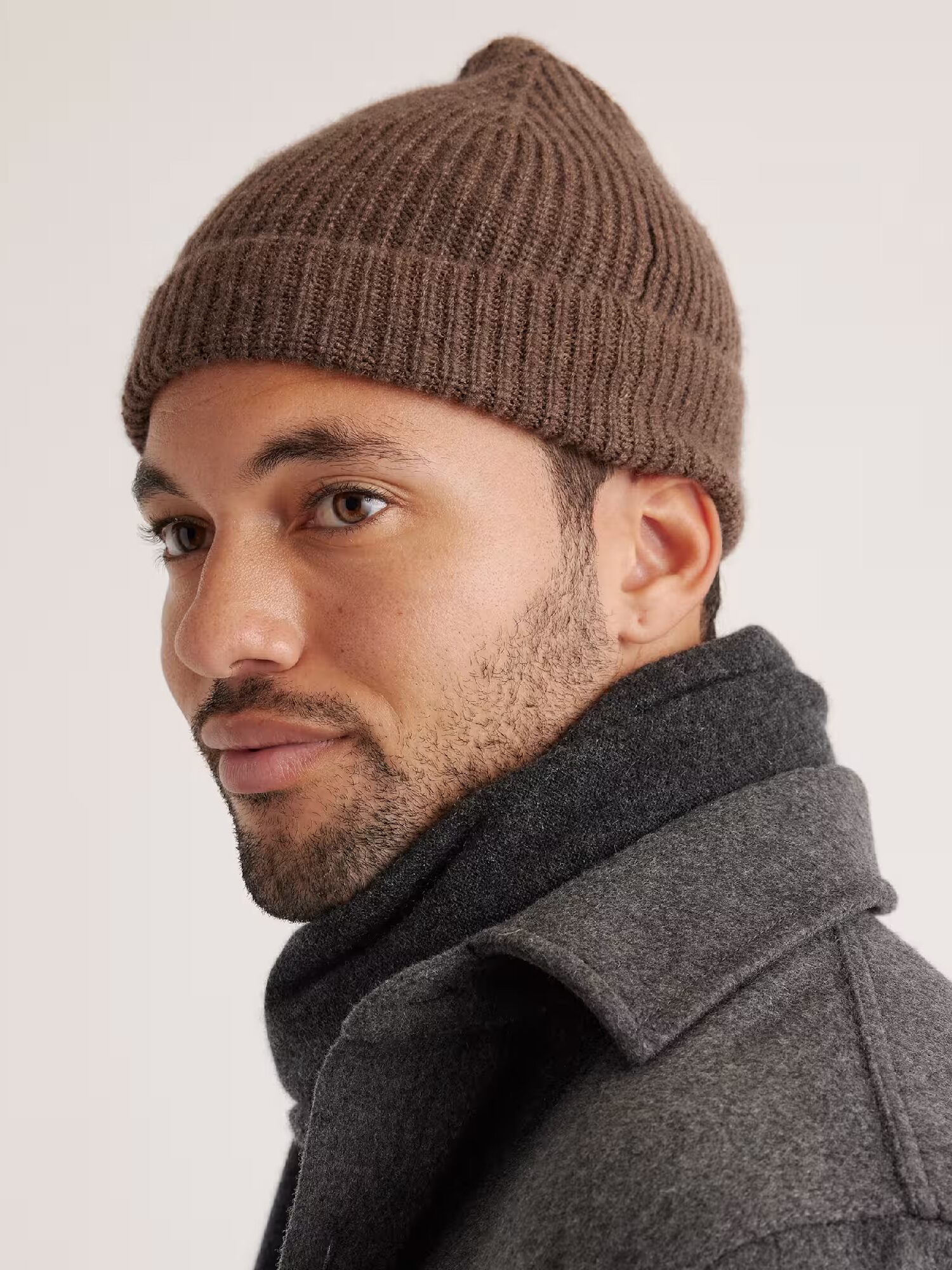 A man wearing a brown knit beanie, a dark scarf, and a grey coat looks to the side against a plain background.