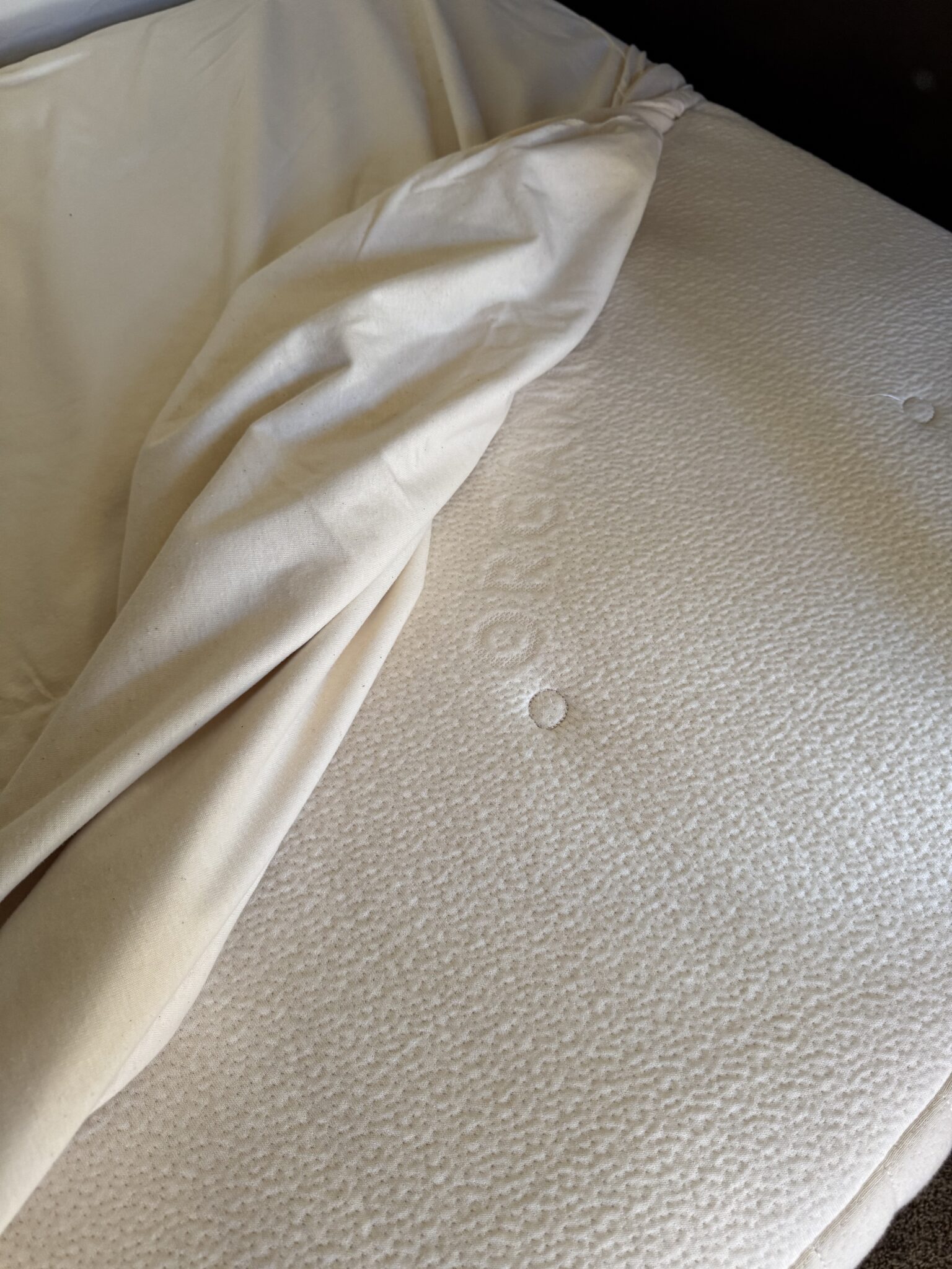 A cream-colored fitted sheet is partially pulled back, revealing a textured mattress surface with faint circular indentations and an embossed brand name.
