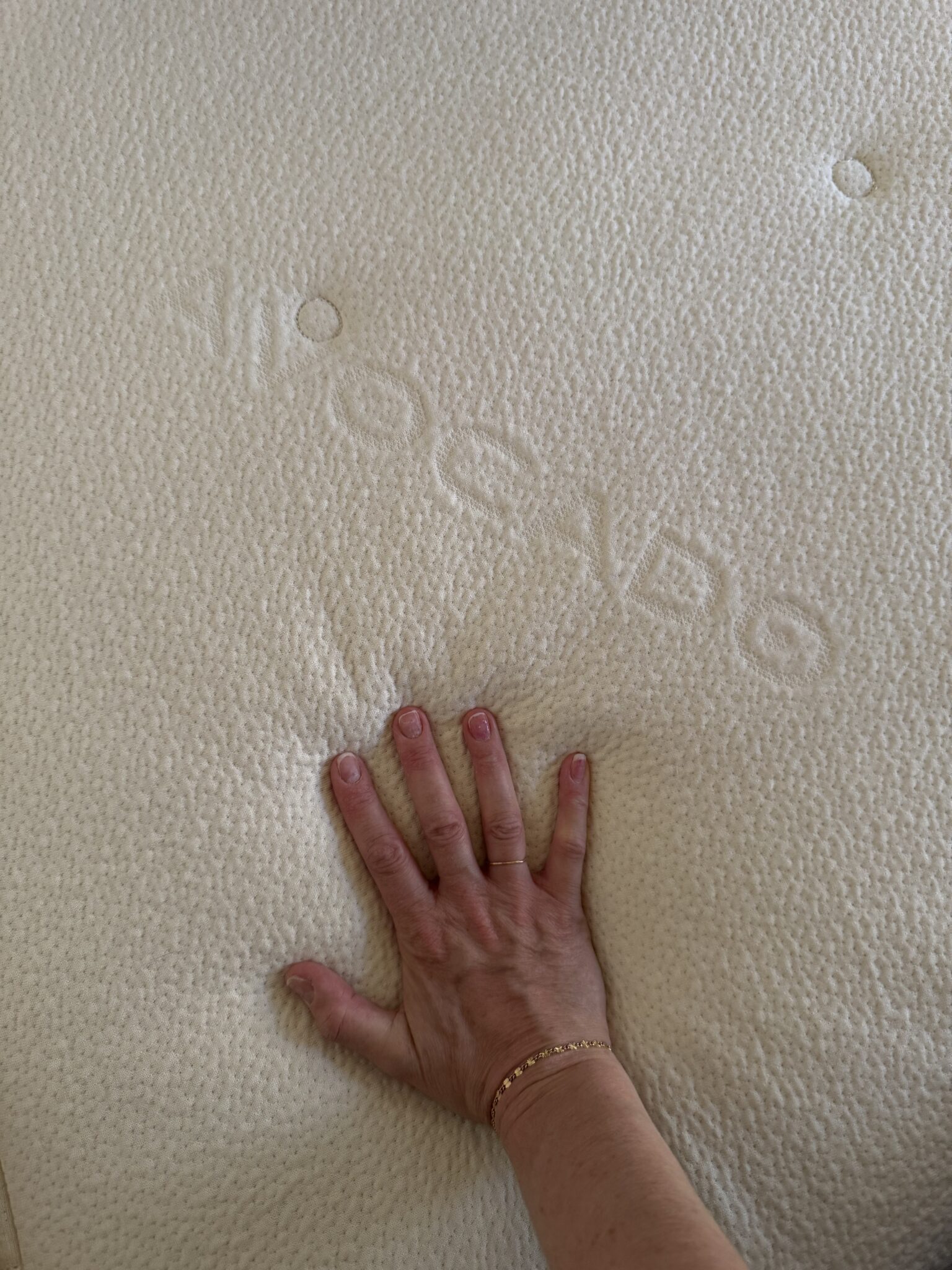 A hand pressing down on a cream-colored mattress, showing an indentation and embossed letters visible on the mattress surface.