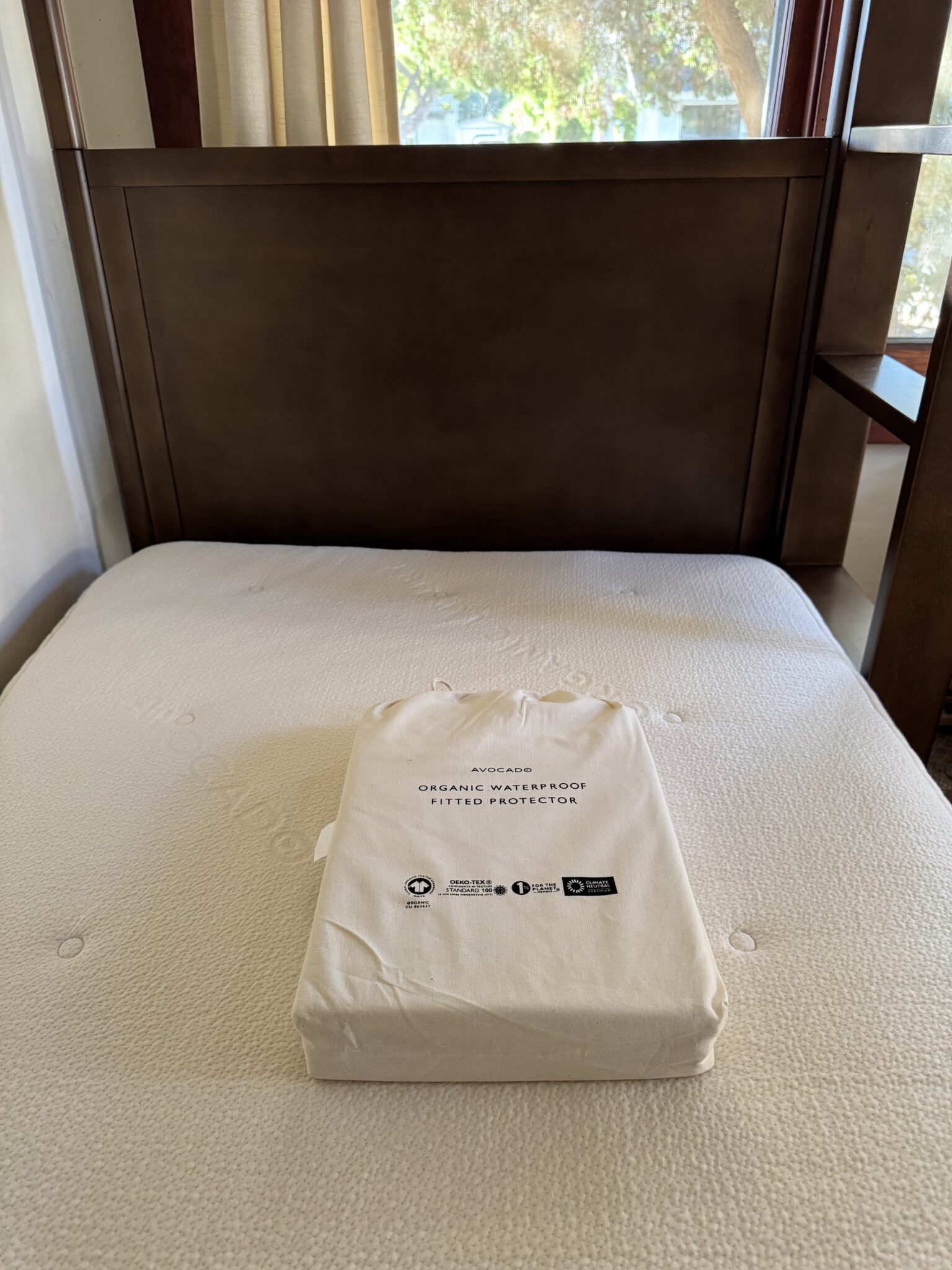 A packaged Avocado brand organic waterproof fitted protector is placed on top of a bare mattress in a wooden bed frame.