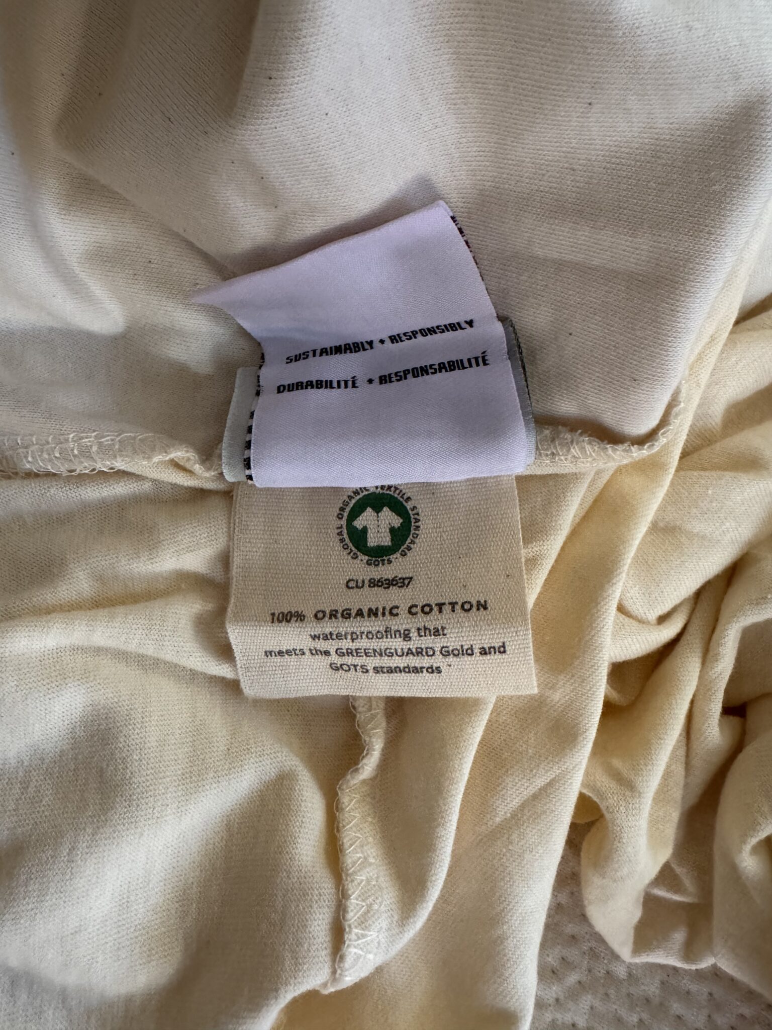 Two clothing tags on a beige cotton fabric; one tag notes sustainability and durability, the other indicates 100% organic cotton and certification details.