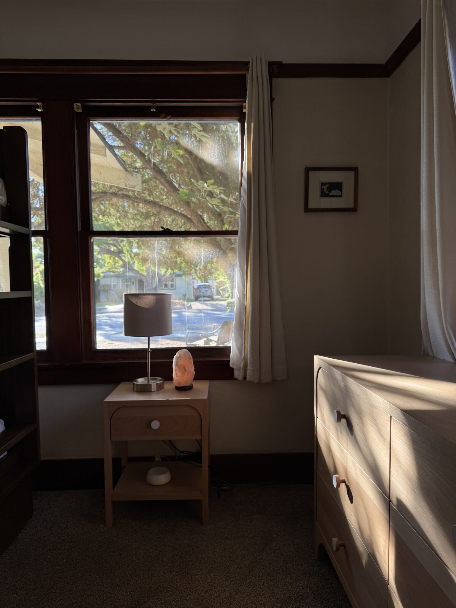 A bedroom corner with a window showing trees outside, a small wooden nightstand with a lamp and salt lamp, and a light wood dresser in natural sunlight.