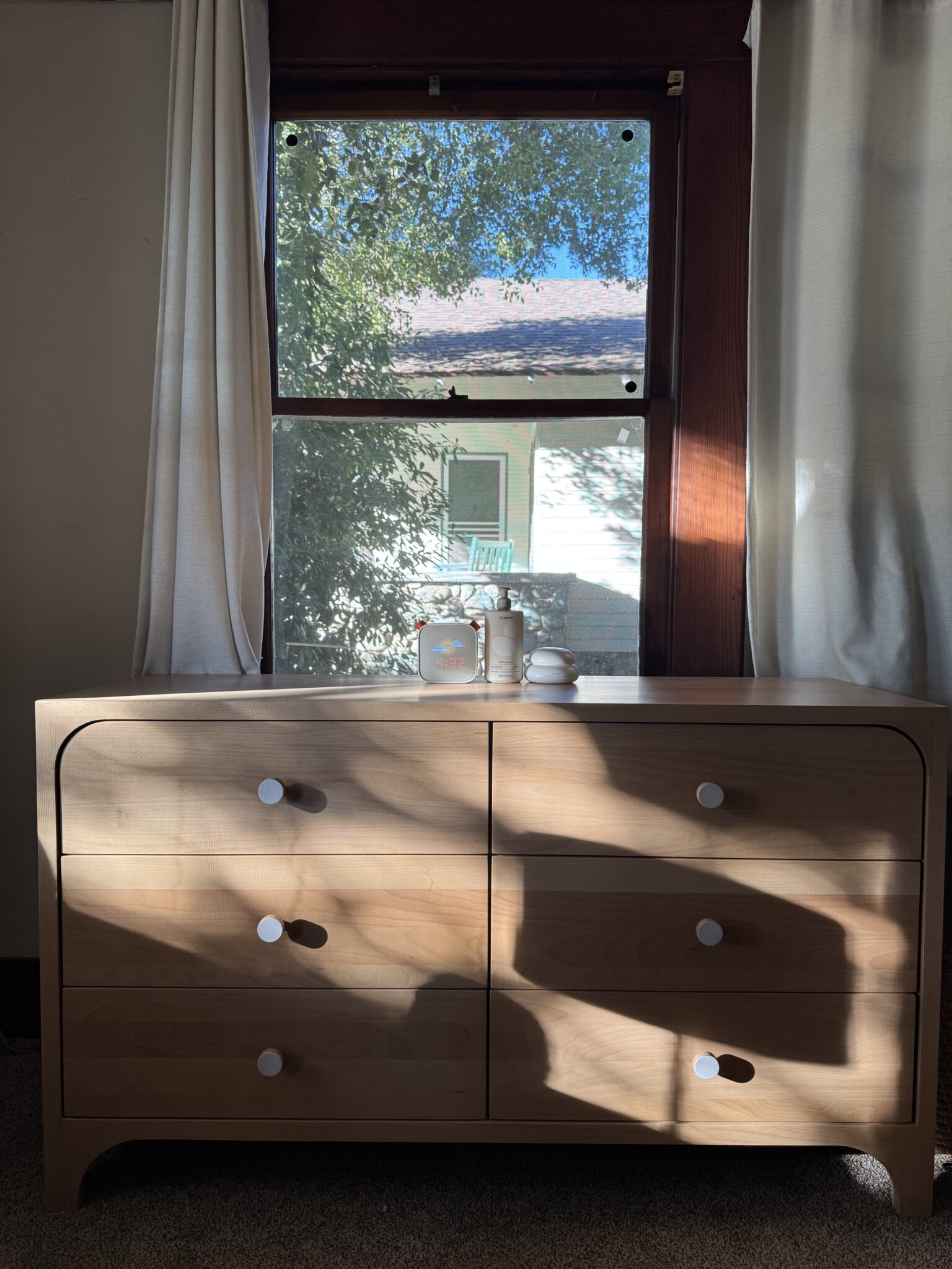 A wooden dresser with six drawers stands in front of a window. Sunlight casts shadows across the dresser. There are a few small decorative items on top of the dresser.