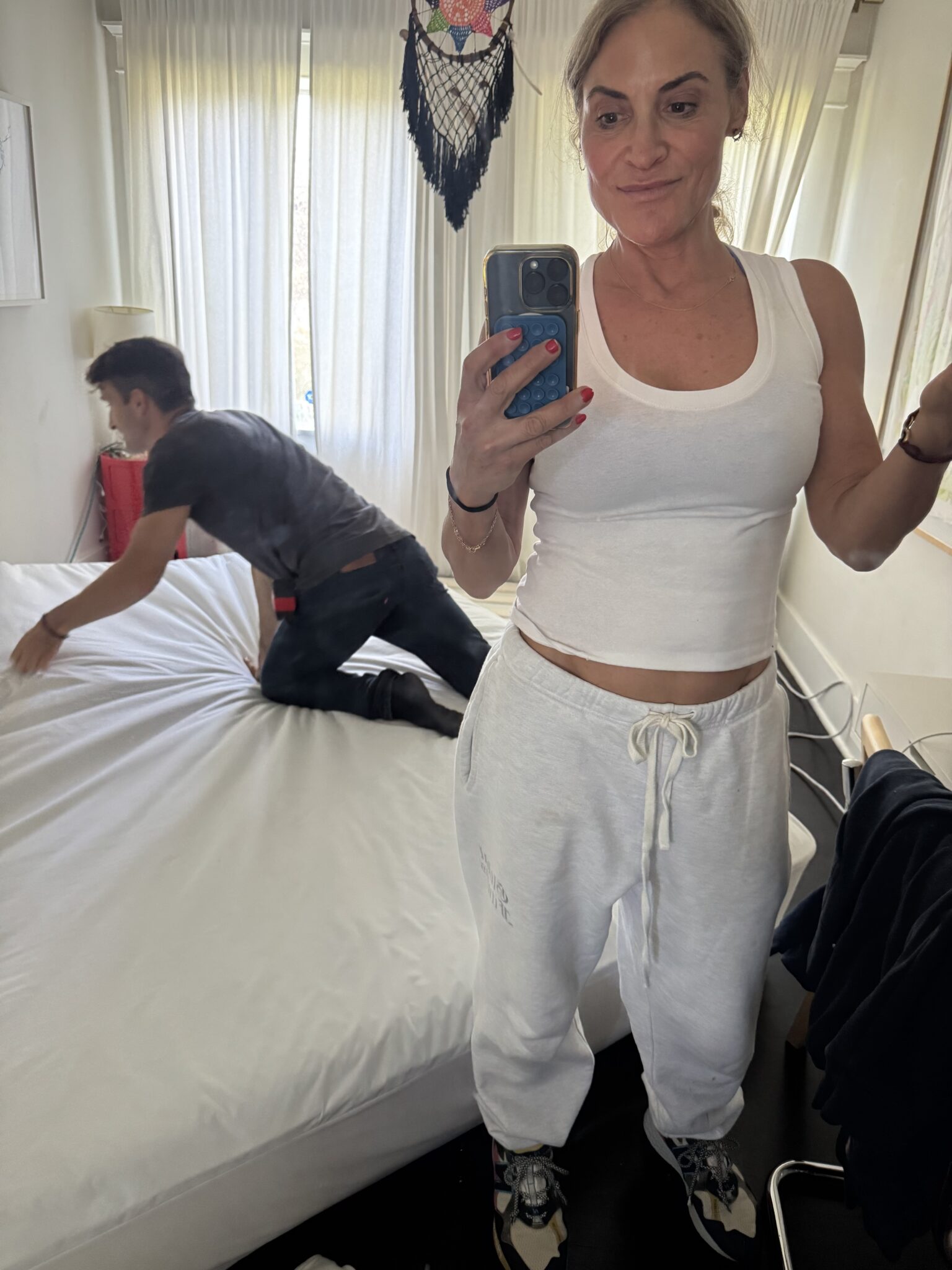 A woman in a white tank top and sweatpants takes a mirror selfie while a man kneels on a bed in the background, making the bed. Natural light comes through sheer curtains.