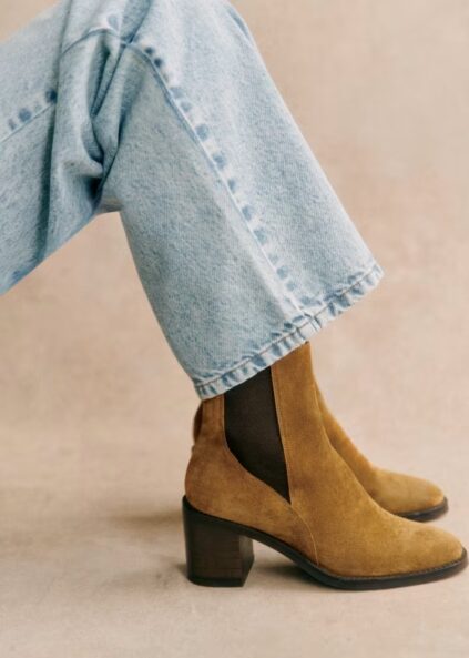 Person wearing light blue jeans and tan suede heeled ankle boots with dark elastic side panels, seated against a neutral background.