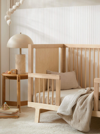 A modern nursery with a light wood crib, beige bedding, a wooden side table with a lamp, toys, and neutral decor on a textured rug.