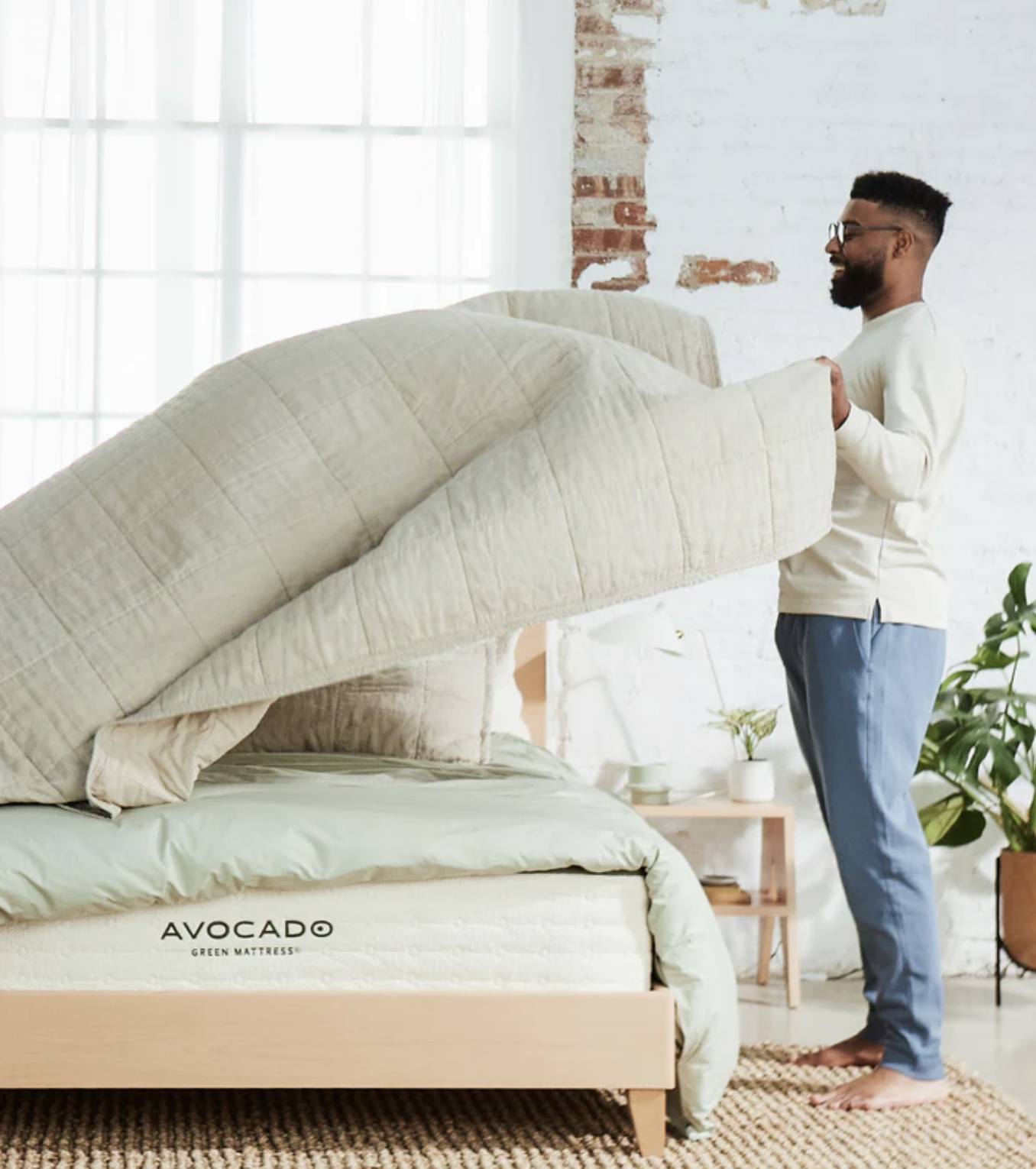 A man stands beside a bed, lifting a light beige comforter. The bed has an "AVOCADO GREEN MATTRESS" label. A plant and small table are in the background.