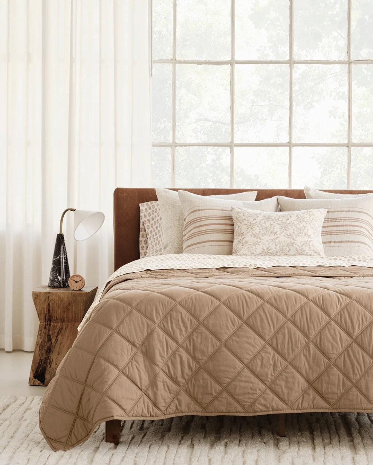 A neatly made bed with tan quilted bedding, multiple patterned pillows, a wooden side table with a lamp and clock, in a bright room with large windows and white curtains.