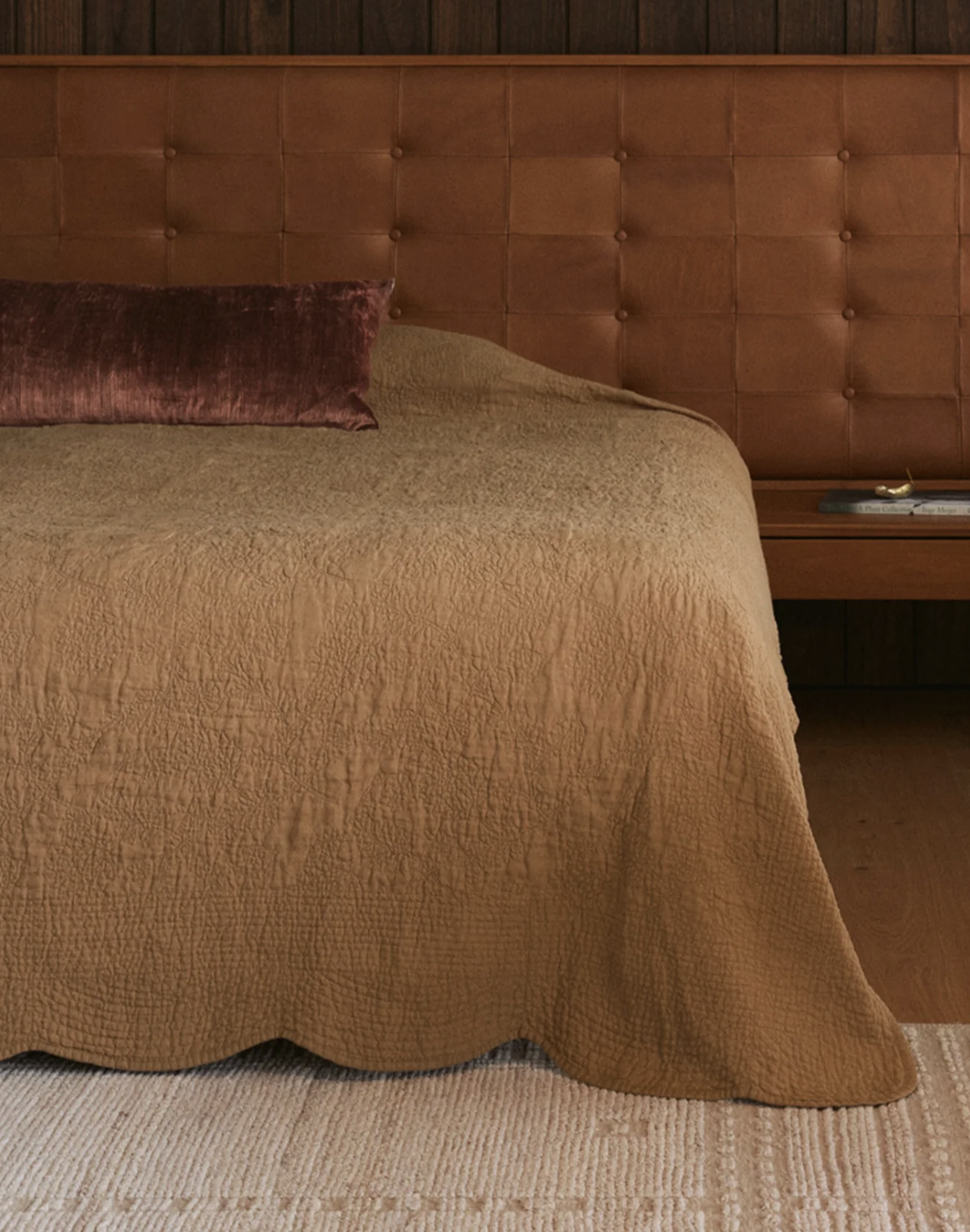 A bed with a brown quilted cover, a tufted leather headboard, a burgundy accent pillow, and a nightstand with a book and small objects.