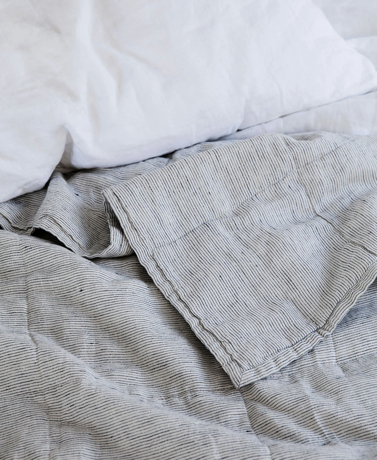 Close-up of a bed with a white pillow and a light gray, finely striped blanket, both appearing slightly rumpled.