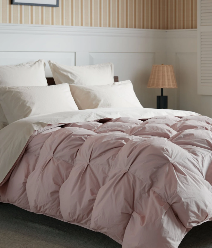 A neatly made bed with white pillows and a light pink, tufted comforter in a bedroom with white paneling and striped wallpaper. A lamp sits on a bedside table.