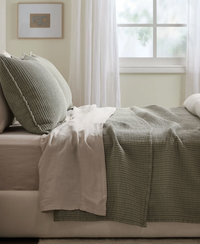 A neatly made bed with textured green bedding and beige sheets sits beside a window with sheer white curtains in a softly lit room.