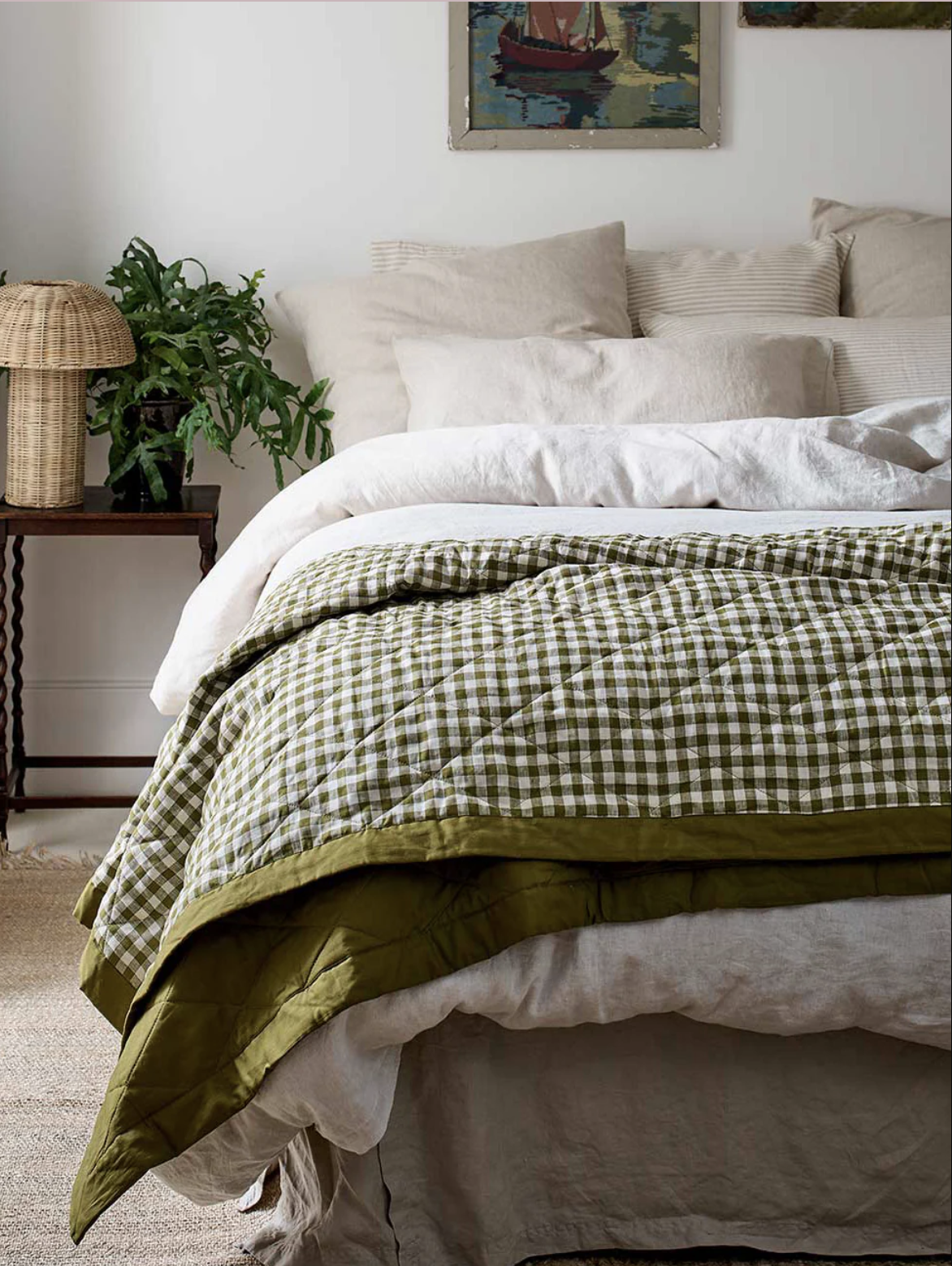 A neatly made bed with white sheets, beige pillows, and a green gingham comforter; a side table with a plant and lamp is next to the bed, and paintings hang on the wall above.
