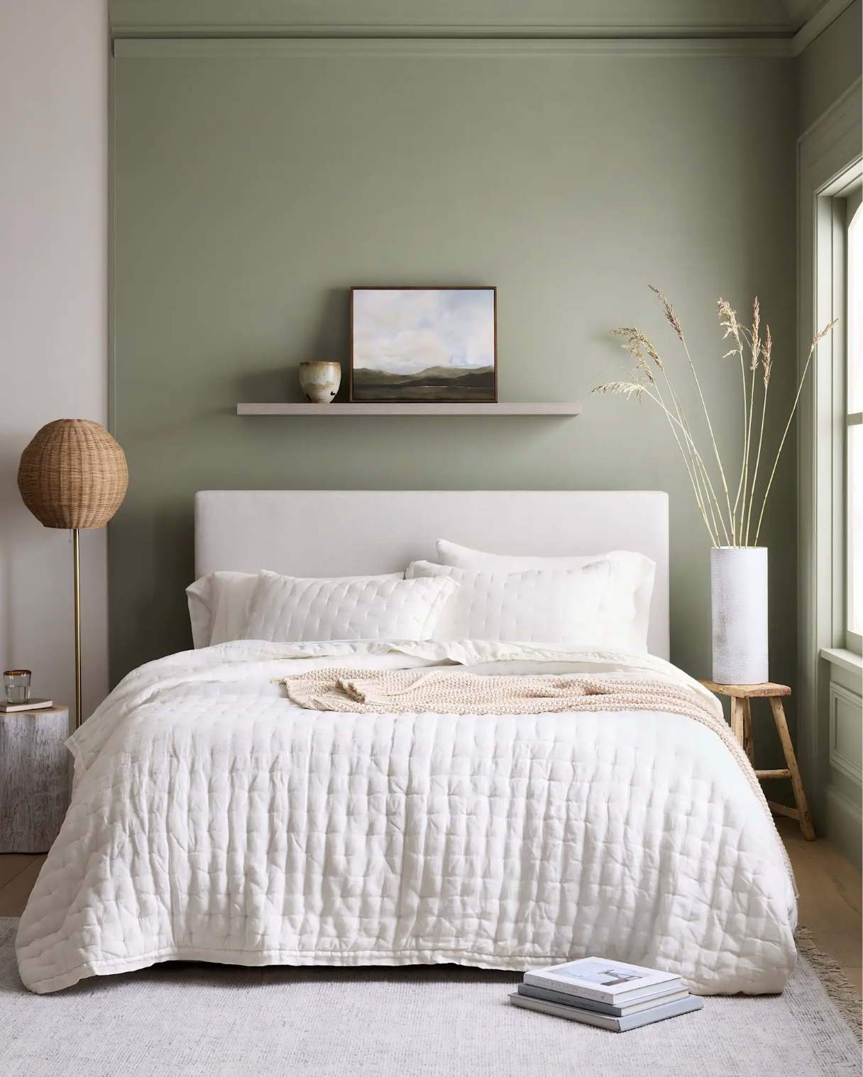 A neatly made bed with a cream quilt, a shelf with decor above the headboard, and a tall vase with dried grass set against a muted green wall in a bright bedroom.