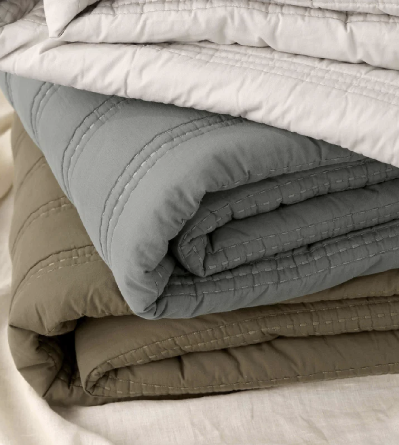 Three neatly folded quilts in light beige, gray, and olive green stacked on a light-colored fabric surface.
