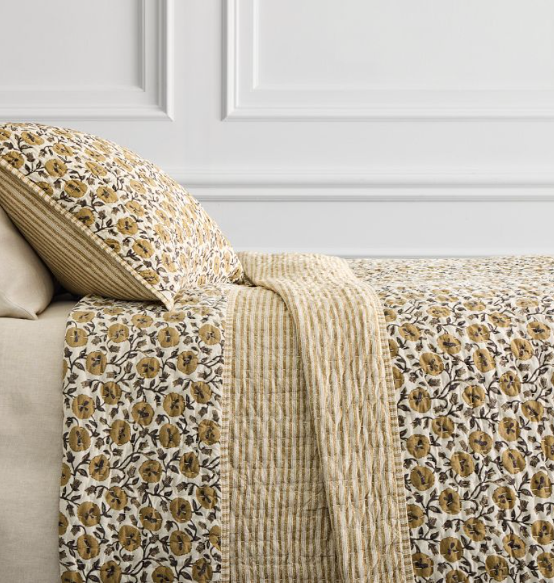 A bed with a beige headboard is made up with a quilt and pillow featuring a yellow and brown floral pattern, accented by beige and yellow striped sections. White paneled wall in background.