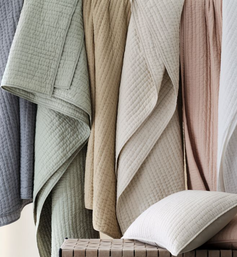 Quilted blankets in pastel shades of green, beige, cream, and pink hang beside a matching pillow on a wooden bench.