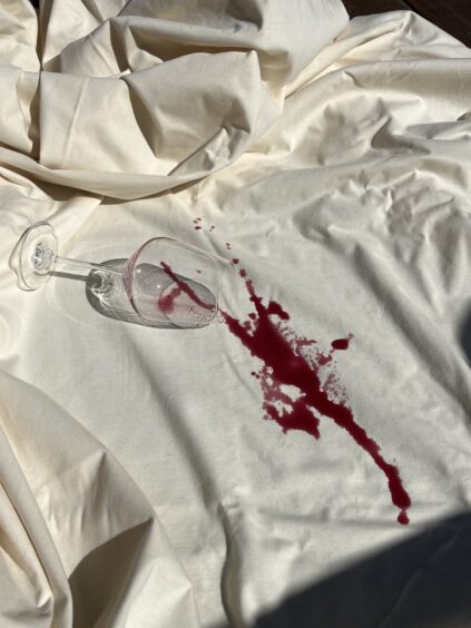 A tipped-over wine glass with red wine spilled onto a white sheet, creating a noticeable stain.
