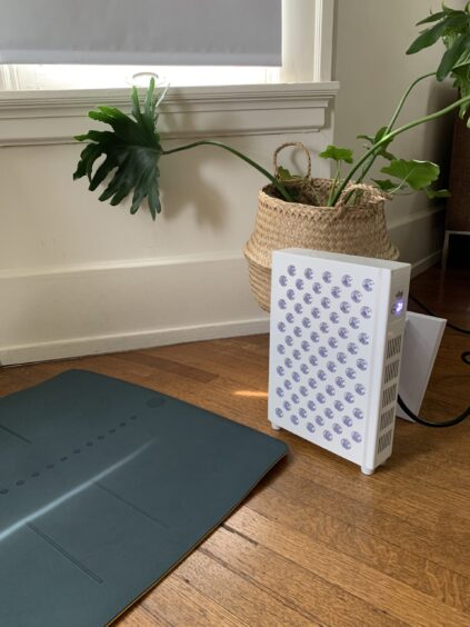 A white LED light therapy panel is plugged in and positioned on a wooden floor next to a woven basket with a green plant. A dark mat lies nearby.