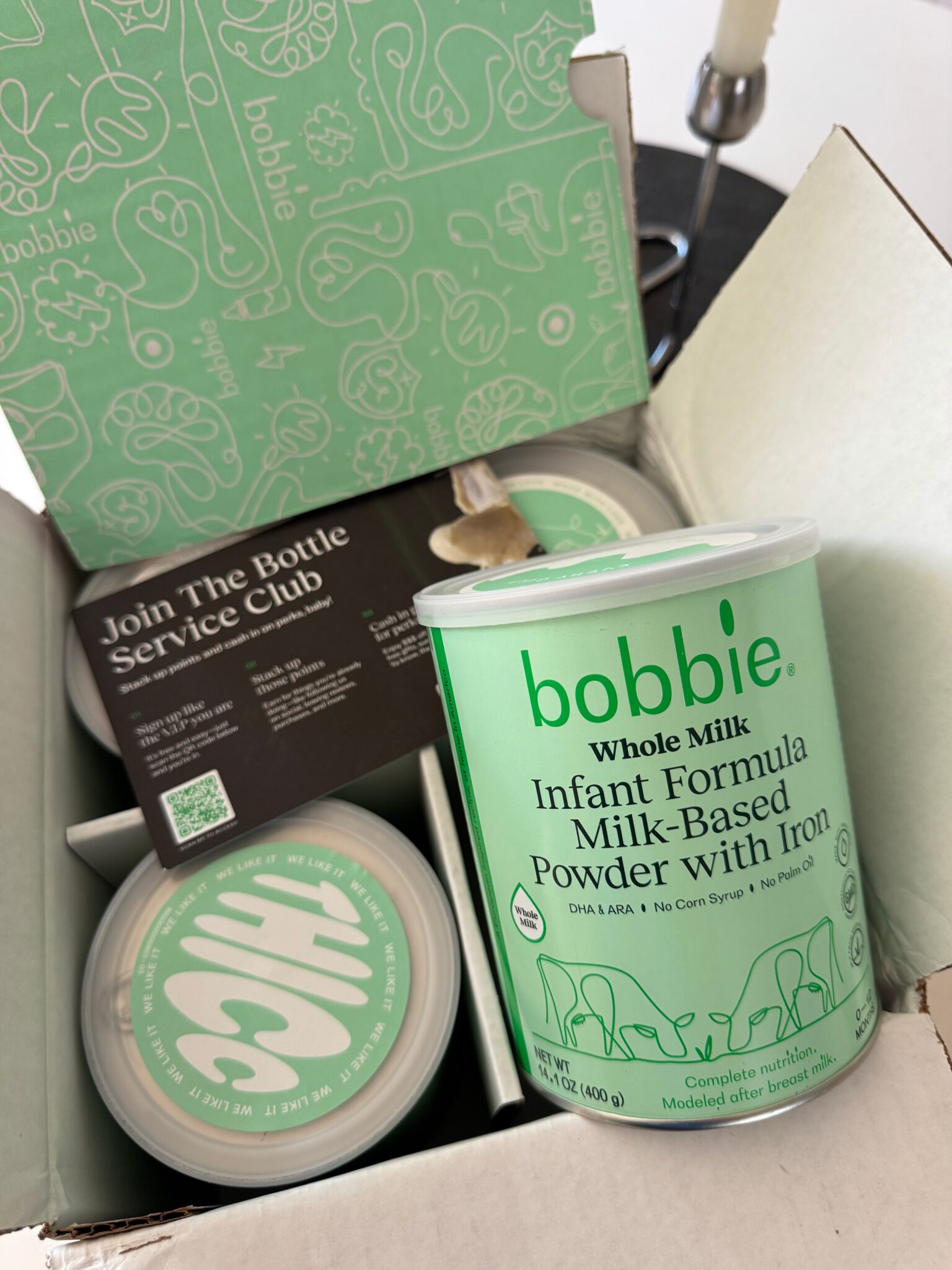 An open box containing cans of Bobbie Whole Milk Infant Formula Powder and informational card, with the packaging featuring light green branding.