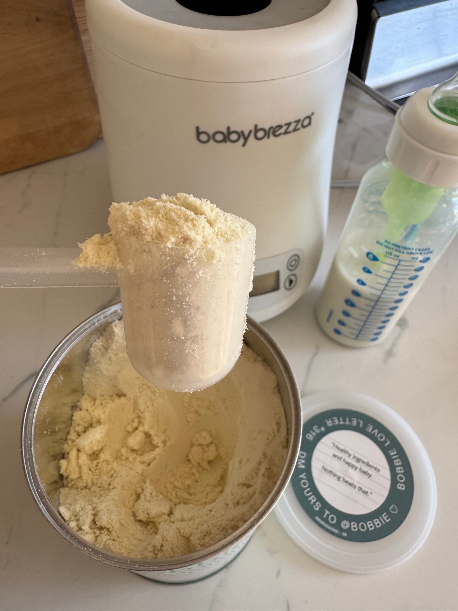 A scoop of powdered infant formula held over an open container, with a Baby Brezza machine and a prepared baby bottle in the background.